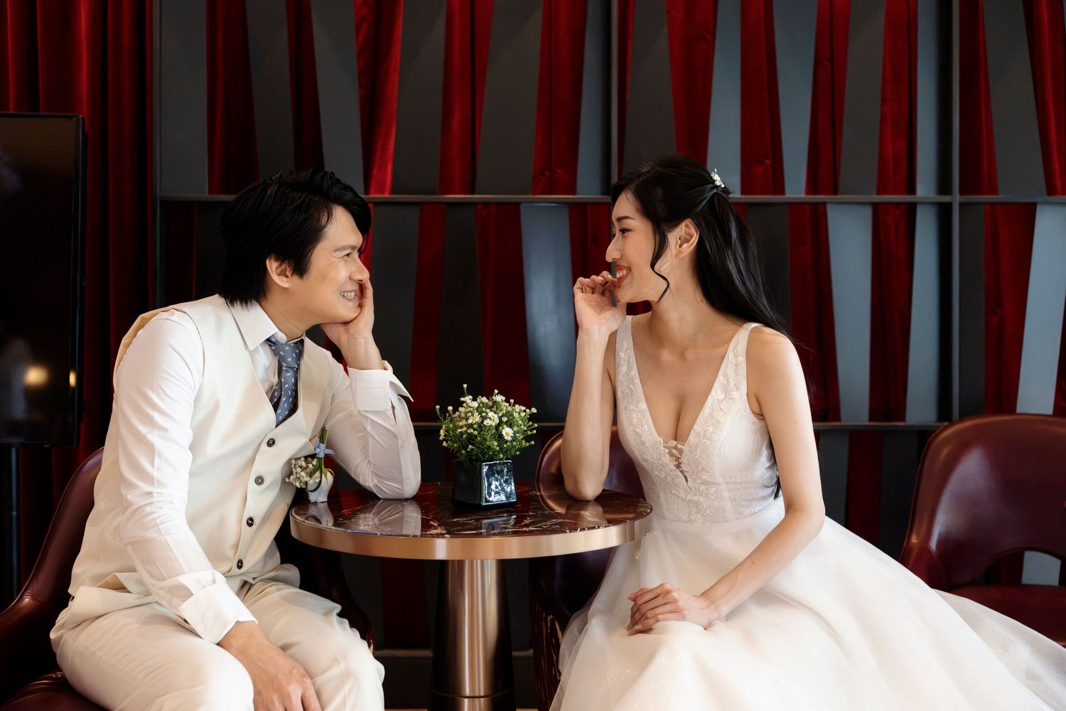 A couple in wedding attire sits at a small table, sharing a romantic moment in an elegant red-curtained venue.