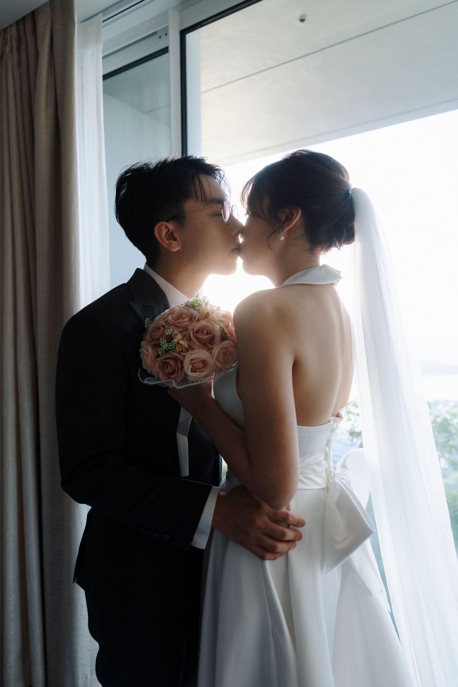 Groom in a black suit kisses his bride in a white halter gown and veil by a sunlit window, holding a pink rose bouquet.