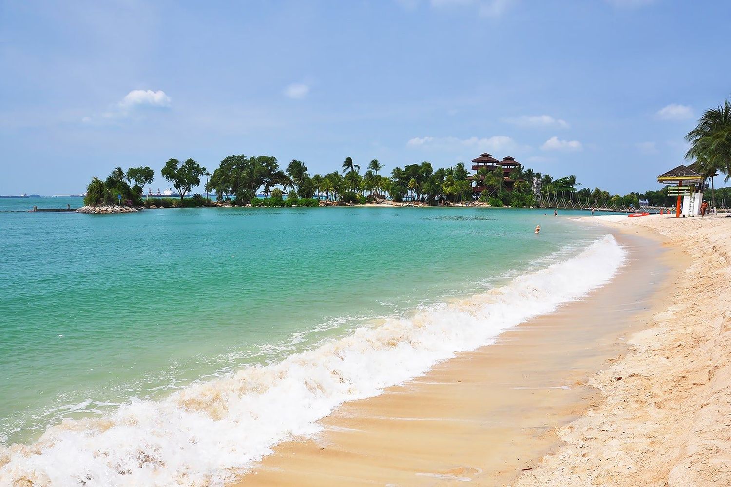 Tropical beach with turquoise waters, golden sand, and lush palm tree island in the background under a blue sky.