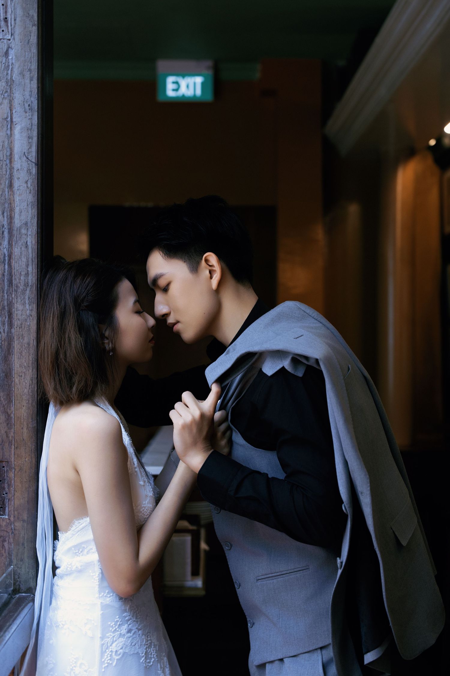 A romantic couple in elegant attire sharing an intimate moment near an exit sign in a dimly lit venue.
