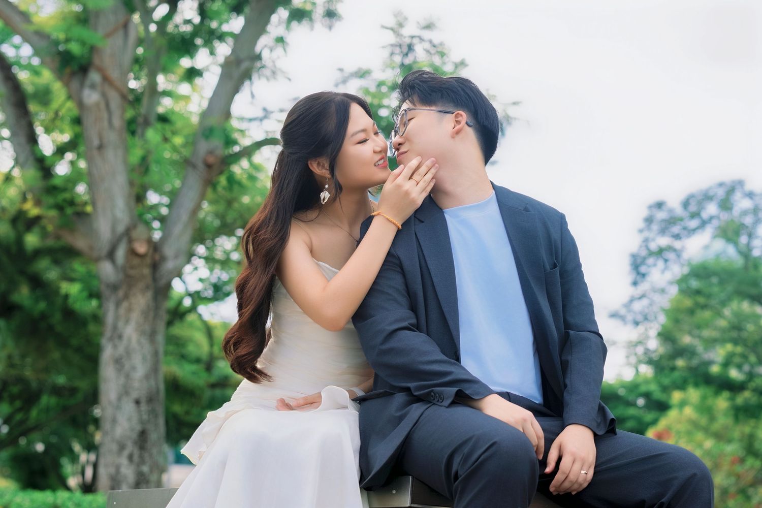 Couple sharing a tender kiss while sitting outdoors surrounded by lush green trees in a park setting.