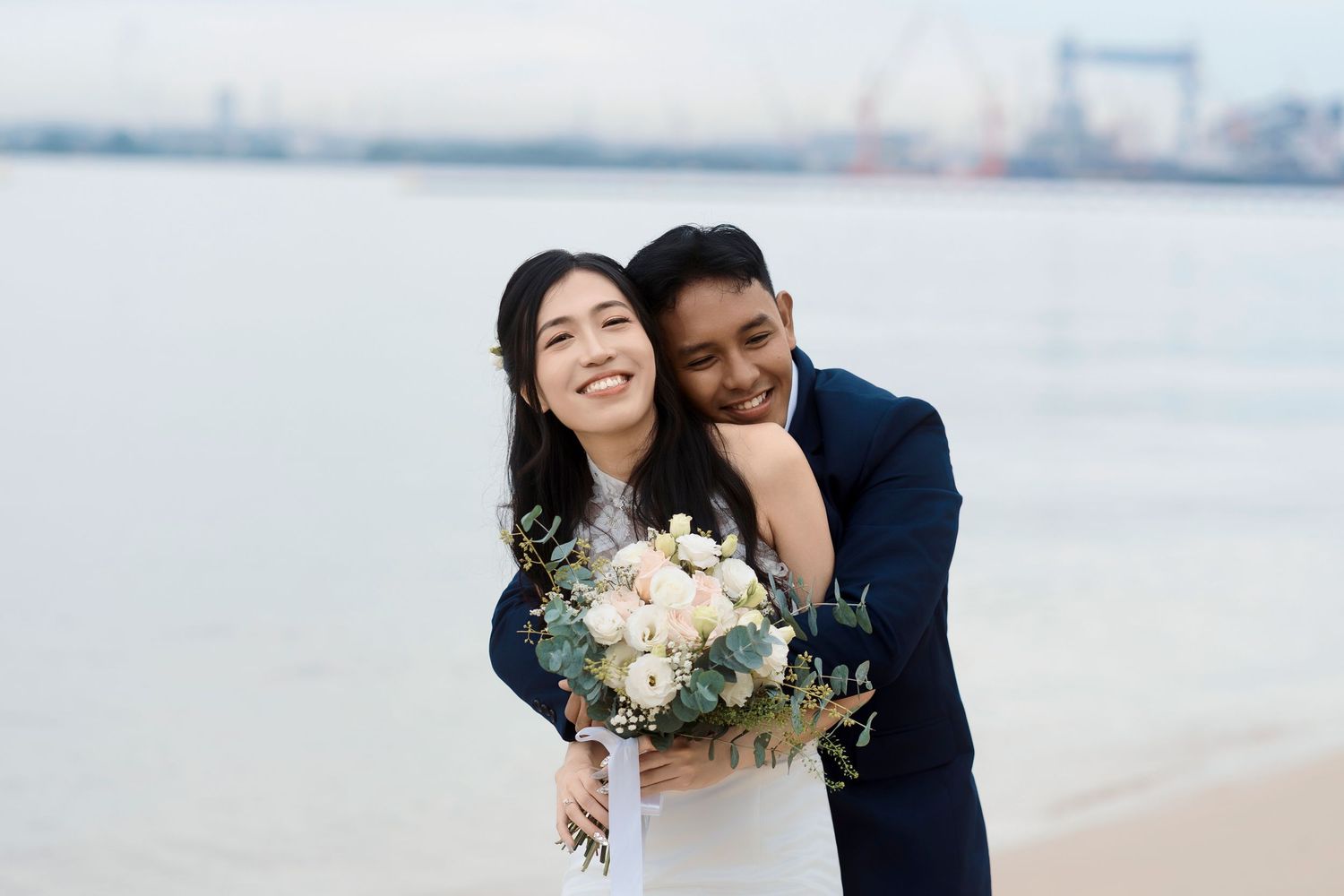 Smiling Asian bride holding white bouquet embraced by groom on a beach with city skyline in the background.