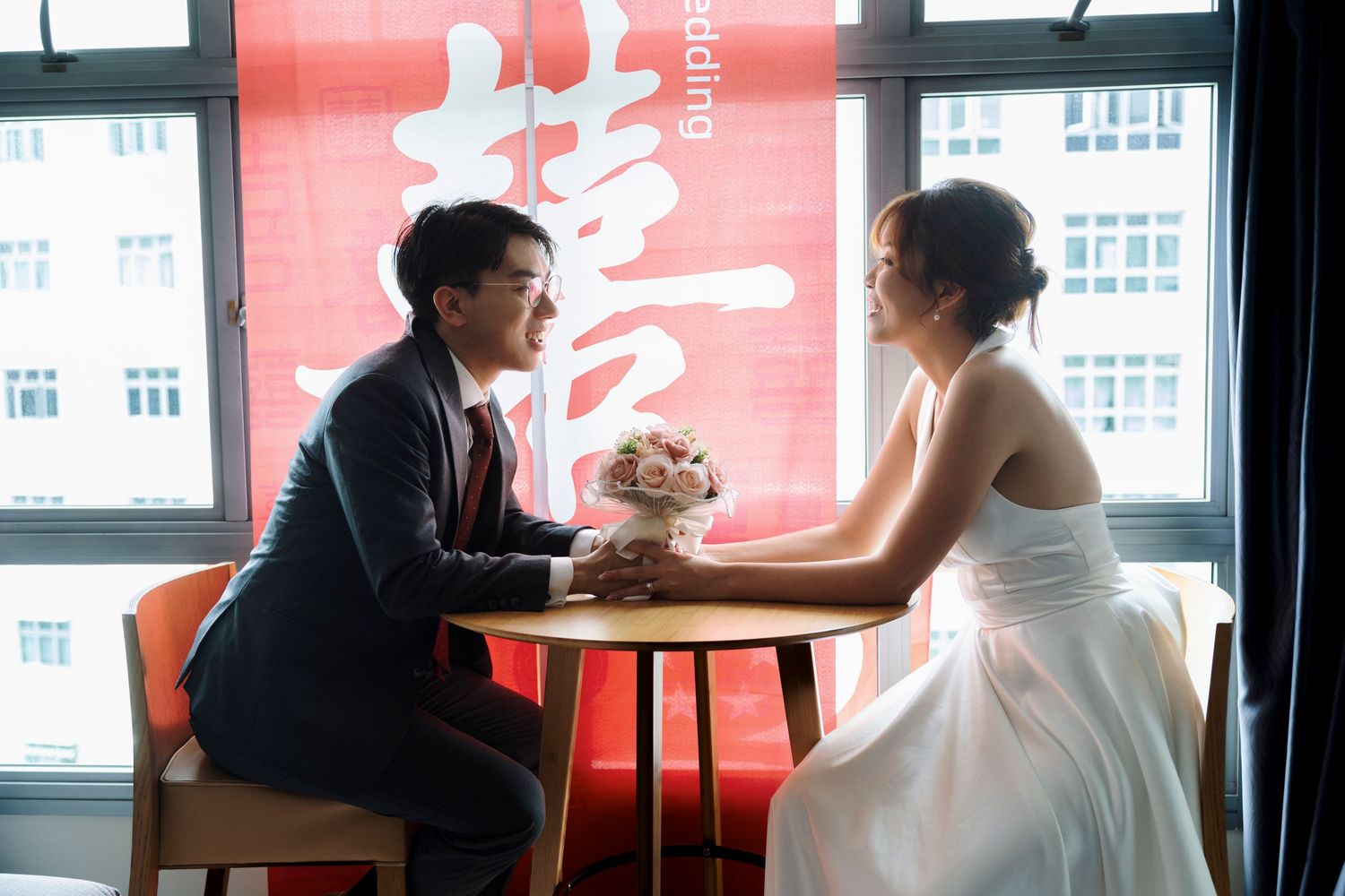Bride in white gown and groom in dark suit sit holding hands at a small table with a bouquet before a red Chinese double happiness banner.