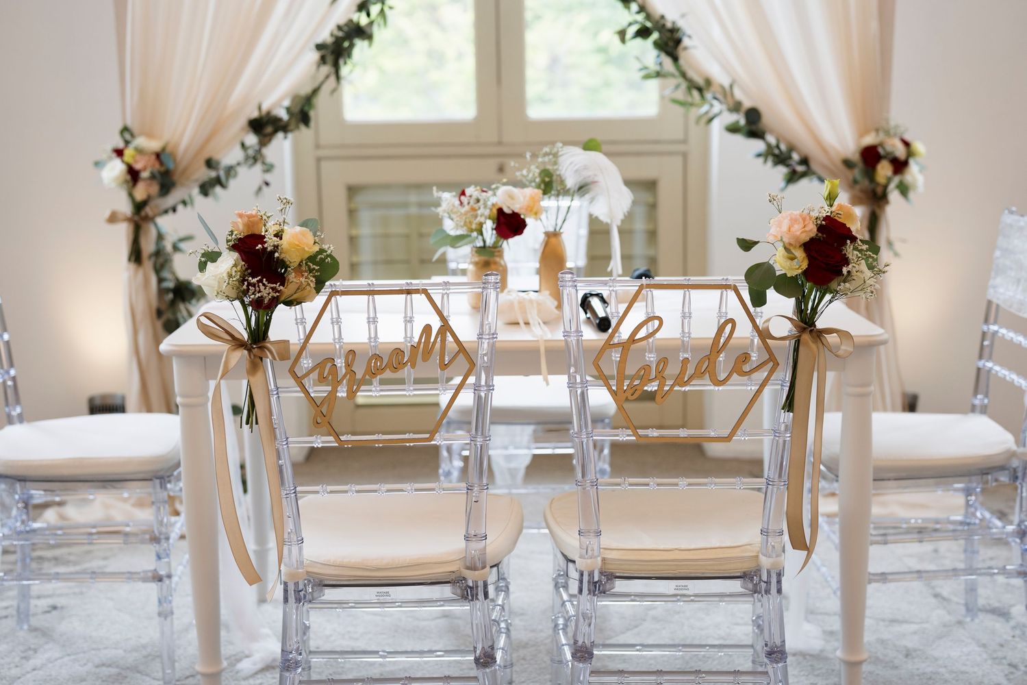 Two clear acrylic wedding chairs with gold script signs and floral bouquets tied with ribbon, set before sheer draped curtains and greenery.