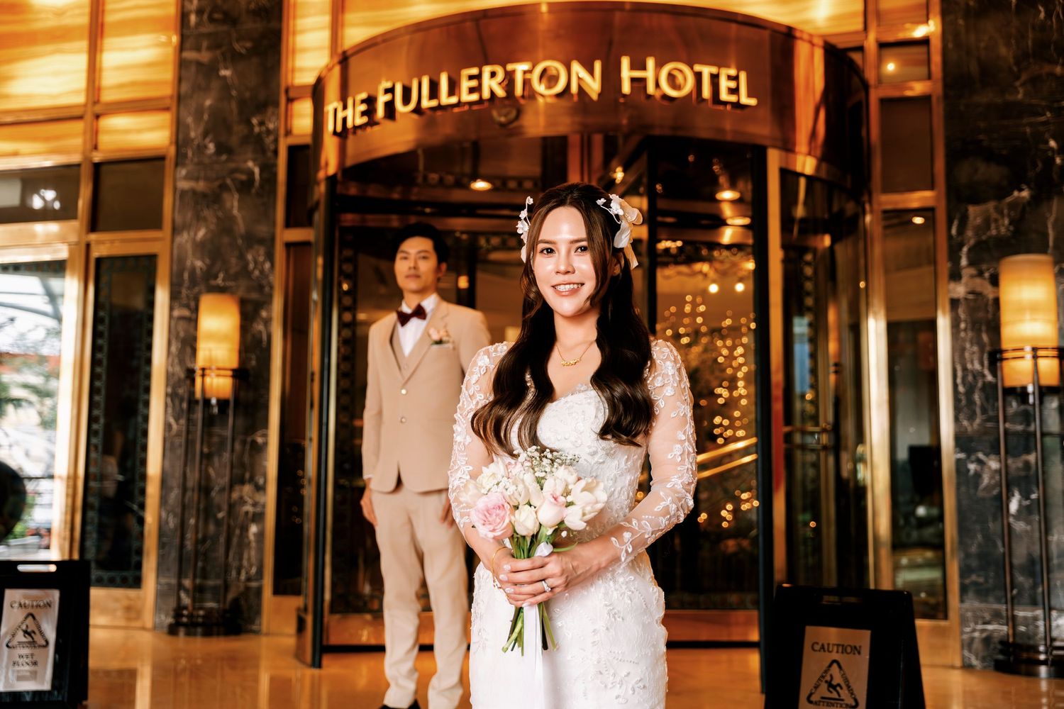 Bride in lace gown holds bouquet outside The Fullerton Hotel entrance with groom in beige suit behind her.