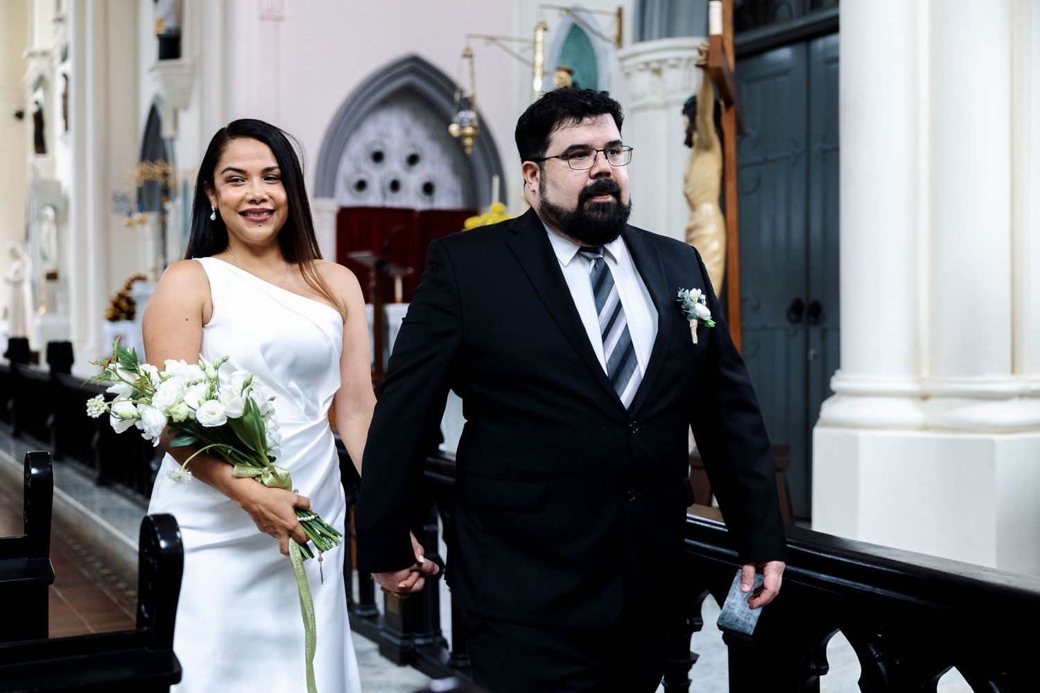 Bride and groom walking down church aisle, bride holding white bouquet in elegant white dress, groom in black suit.