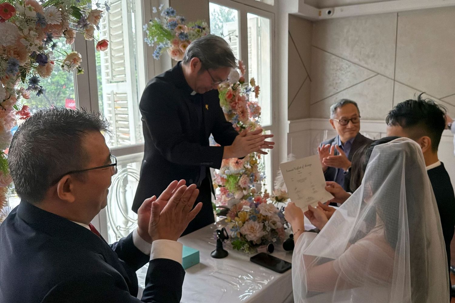 Wedding ceremony with officiant reading vows as bride in white veil and groom in suit stand at floral-decorated altar.