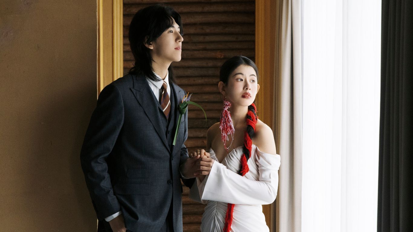 An elegant couple in formal attire standing by a window, man in suit and woman in white dress with red scarf.