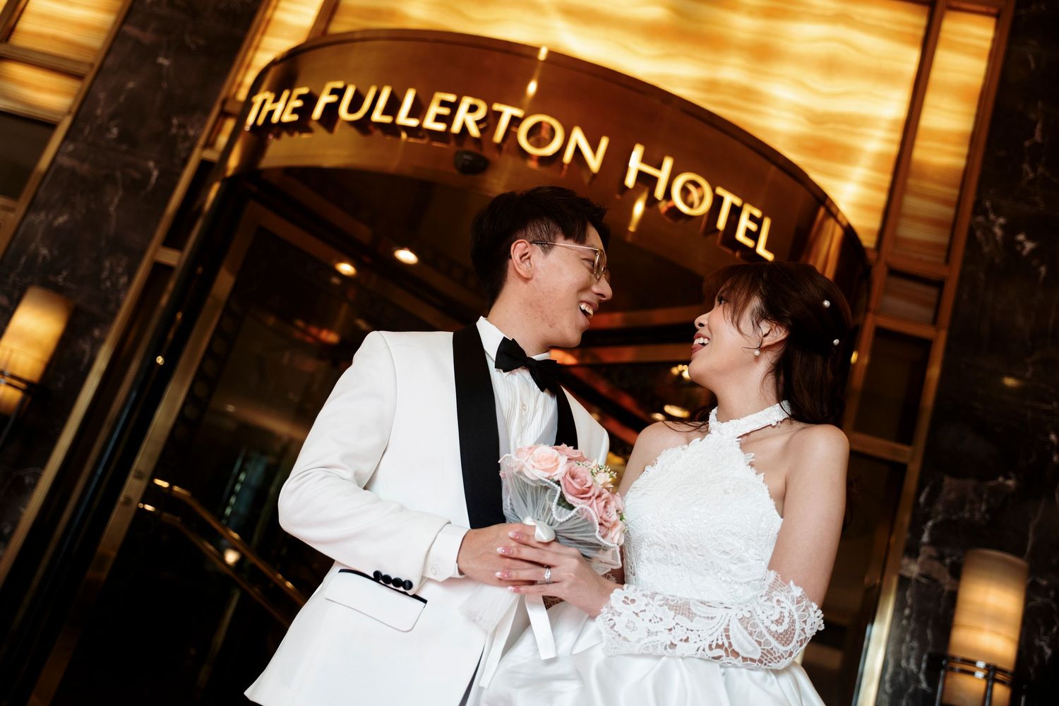 A smiling bride and groom in a white tuxedo jacket pose with a bouquet beneath the illuminated entrance sign of The Fullerton Hotel.