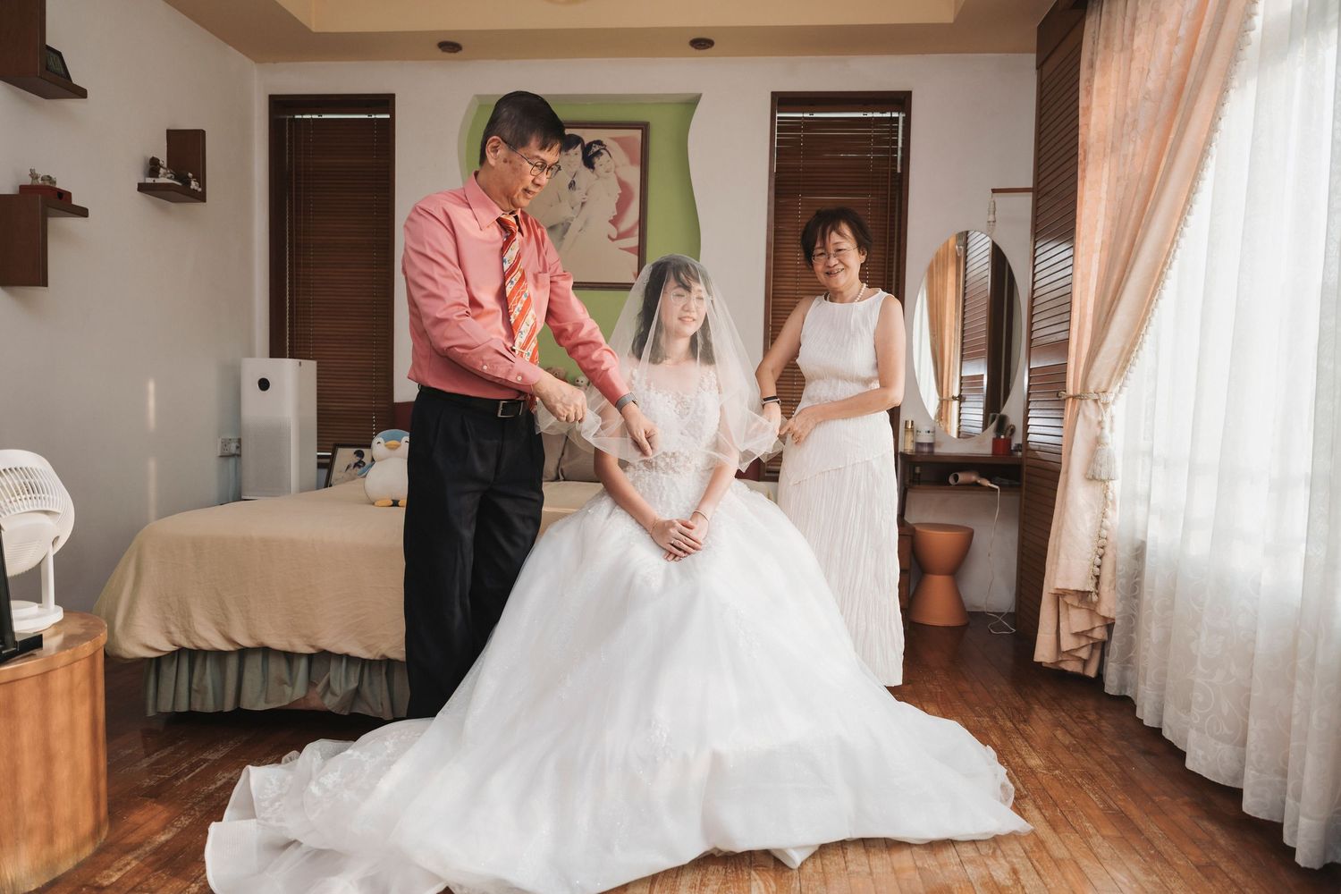 Bride in white wedding gown being helped by family members in a warmly decorated bedroom before the ceremony.
