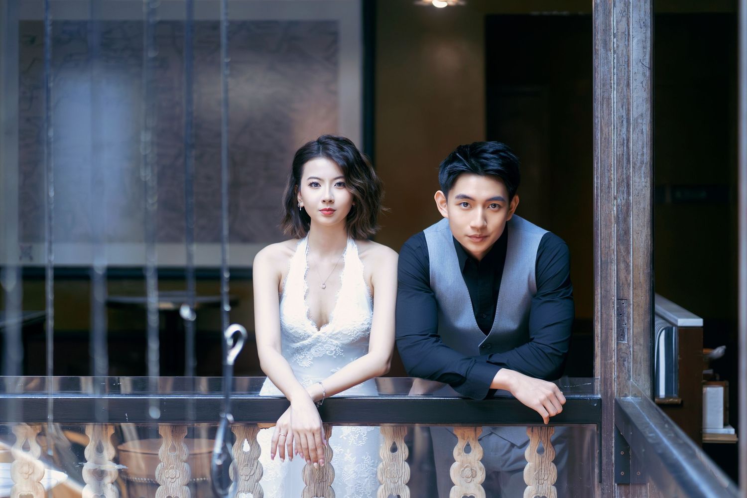 Elegant couple leaning on a balcony railing, woman in white gown and man in dark blazer with moody backdrop.