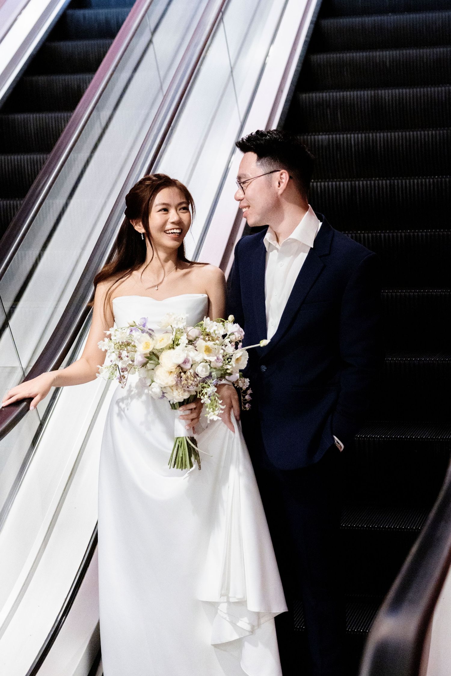 A smiling bride in a white gown holds a bouquet beside her groom in a navy suit on an escalator.