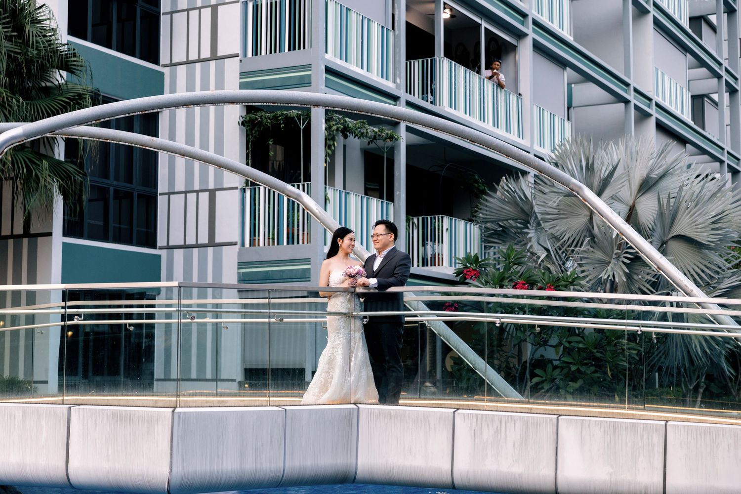 Bride and groom share a romantic moment on a modern bridge with sleek architecture and tropical greenery.
