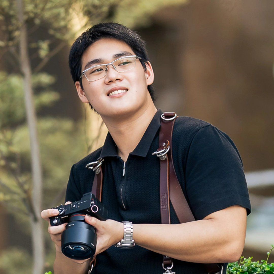 Smiling Asian male photographer holding a camera with leather strap outdoors in natural light.