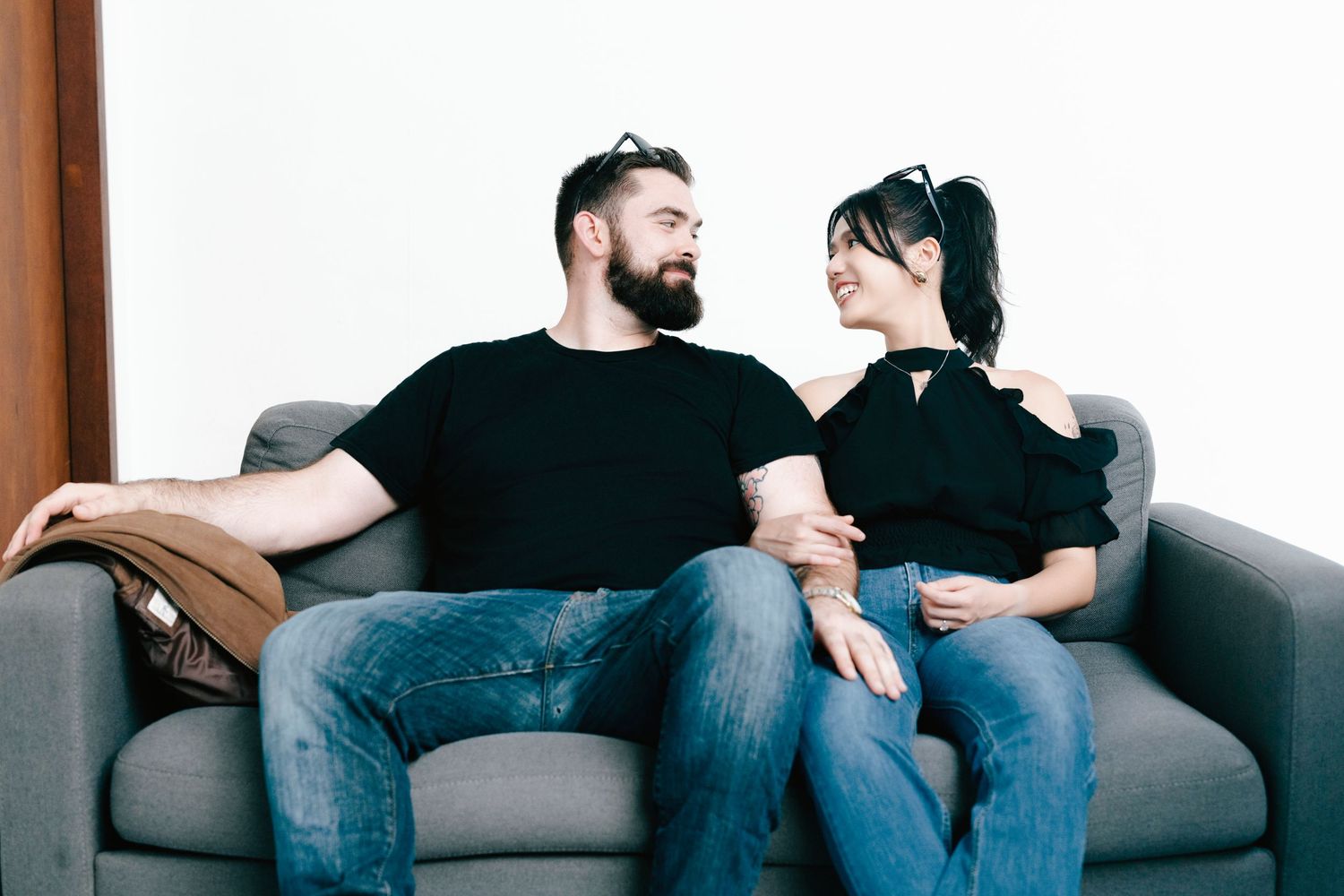 Happy couple sitting on a gray sofa, smiling at each other, wearing black tops and jeans in a bright room.