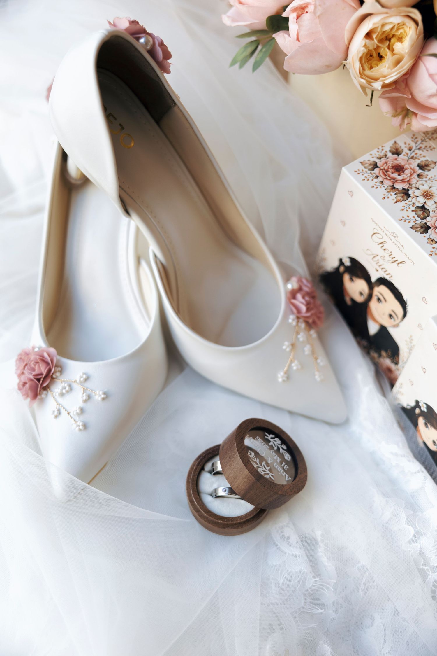 White bridal heels with pink floral accents beside a wooden ring box and pink roses on white lace.