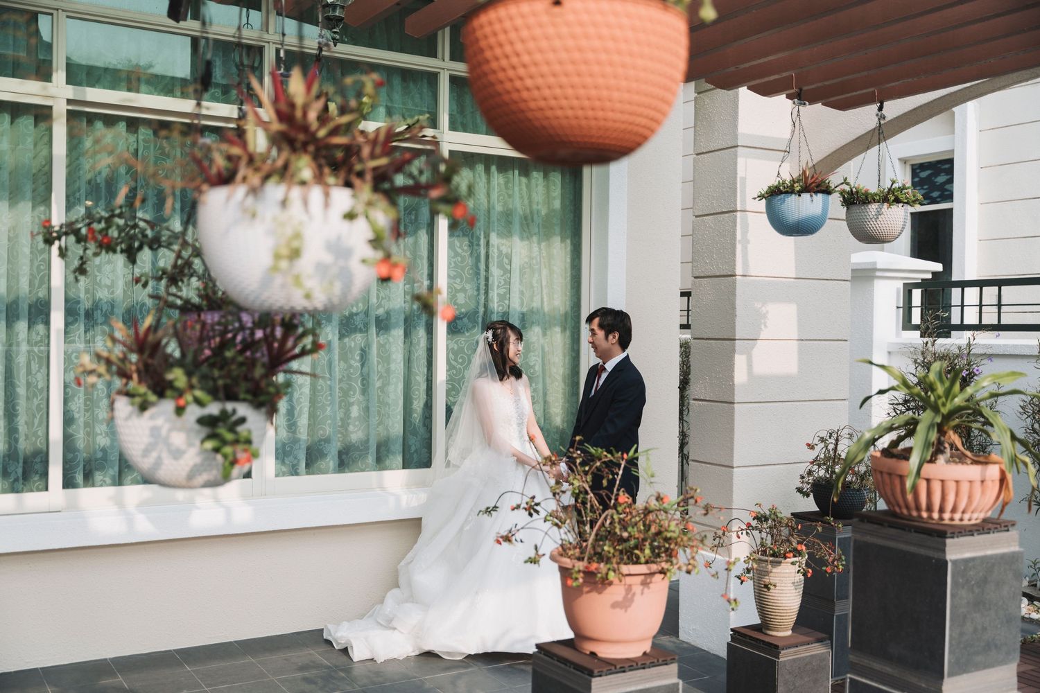 A bride and groom share a romantic moment on a porch surrounded by lush hanging plants and potted flowers.
