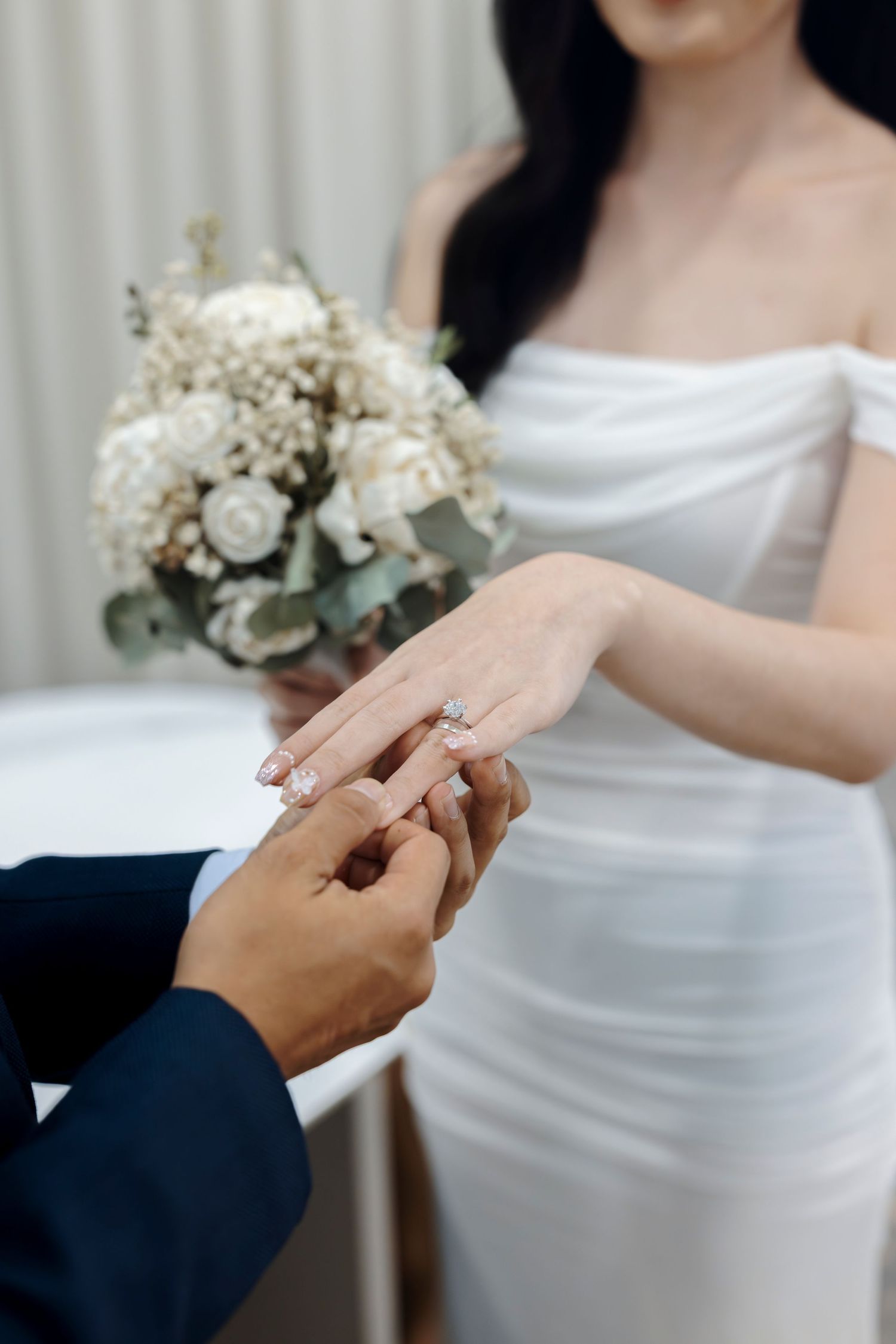Groom placing a diamond ring on bride's finger, bride holding white rose bouquet, wearing off-shoulder wedding dress.