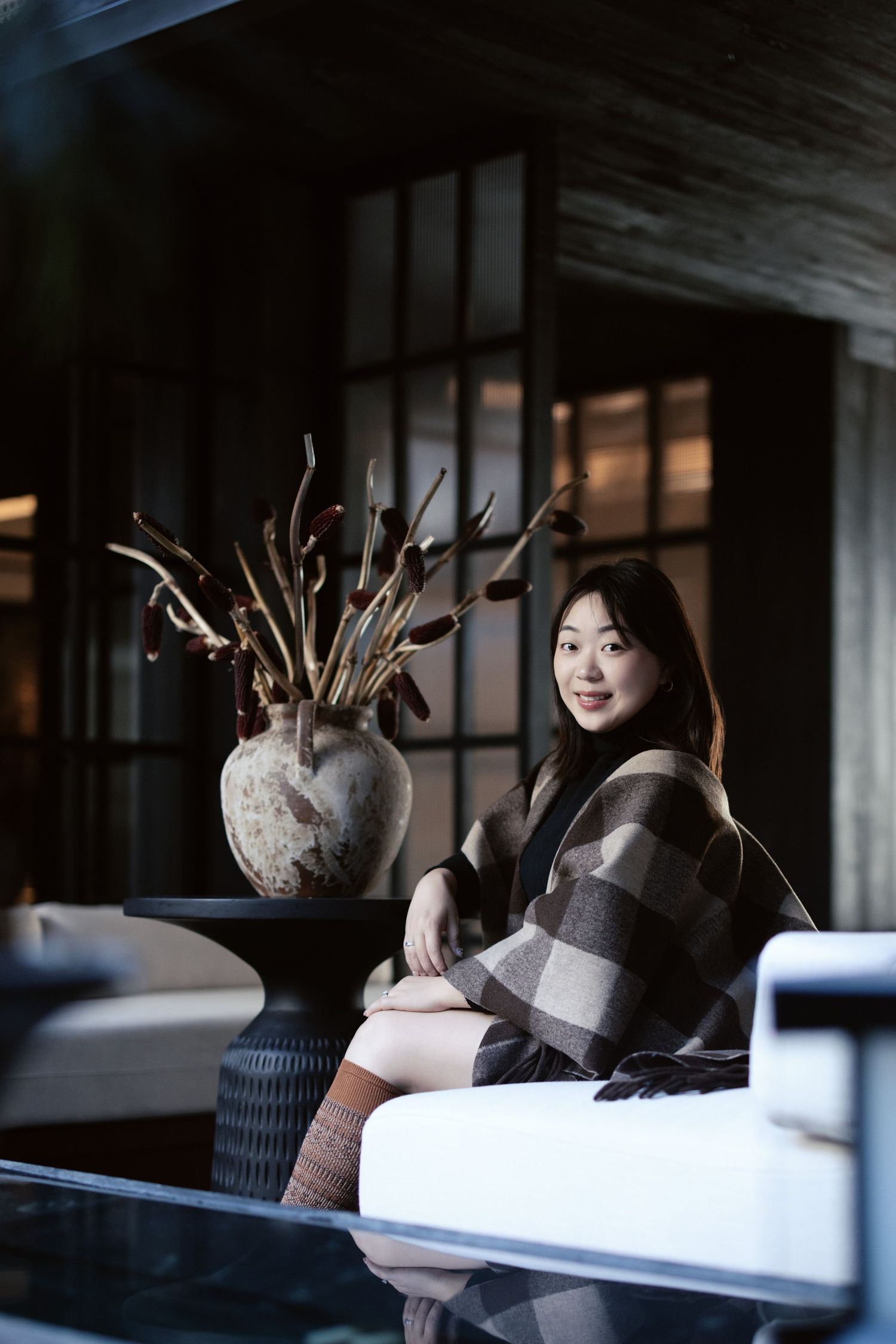 Young Asian woman in plaid wrap sitting beside a decorative vase in a modern dark interior.