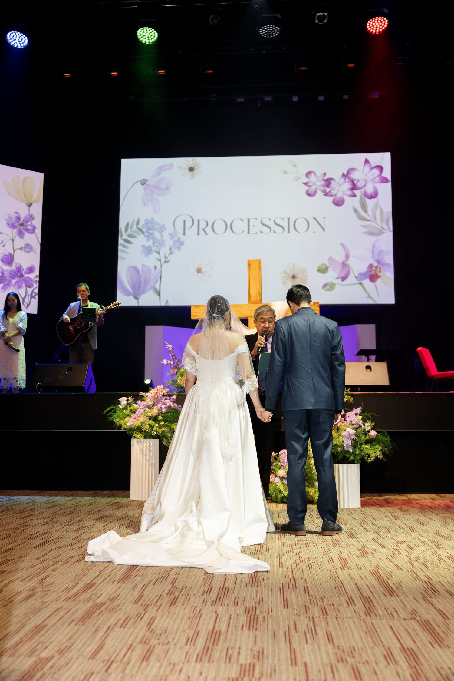 A bride and groom stand before an officiant at a Christian wedding ceremony with a floral Procession screen backdrop.