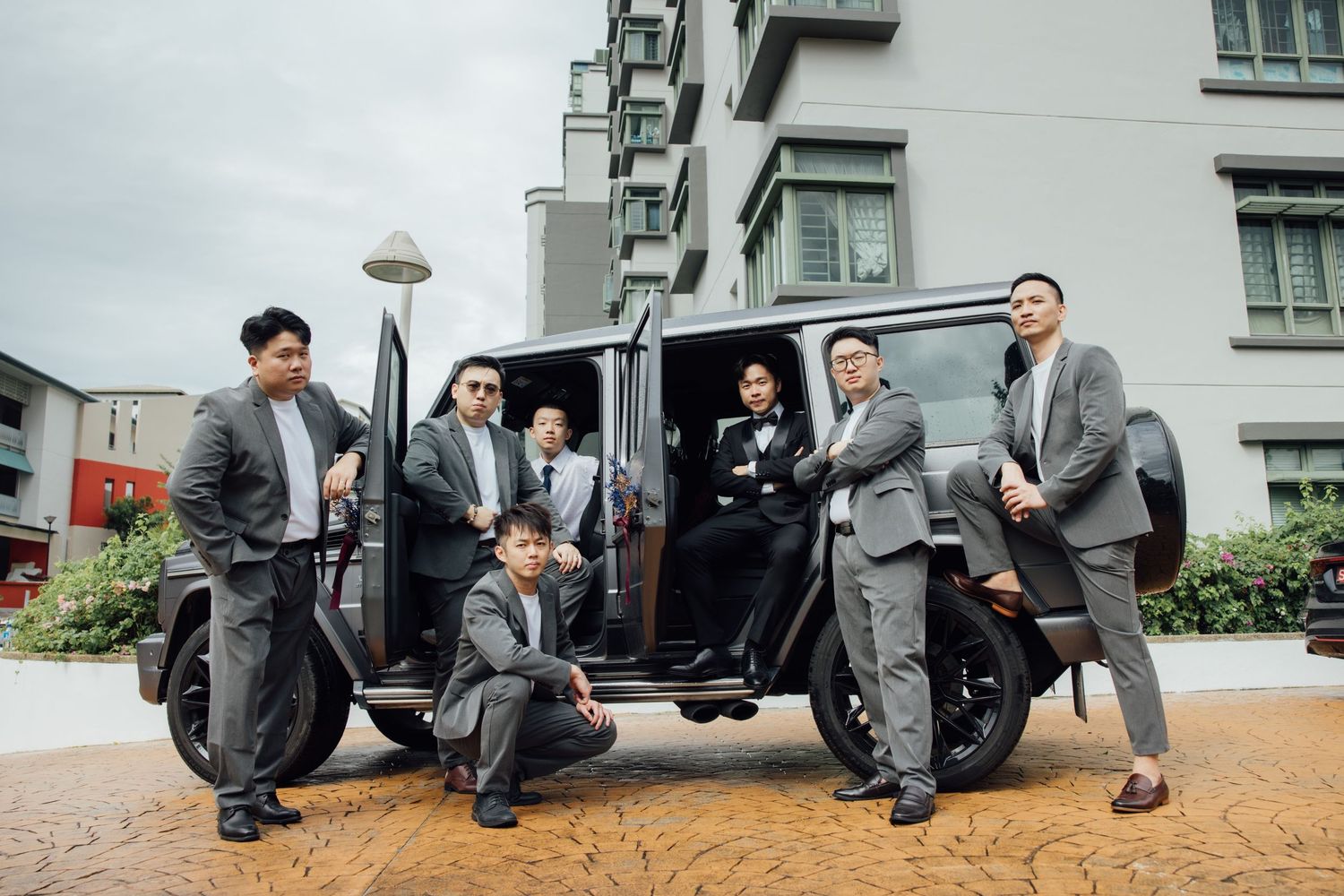 Groomsmen in gray suits pose around an open black Mercedes G-Wagon on a brick-paved courtyard in front of a modern apartment building.