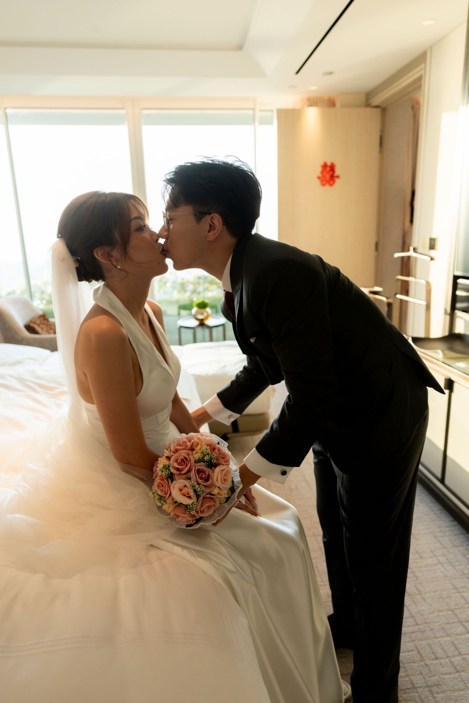 A bride in a white halter wedding gown holding a pink rose bouquet shares a kiss with her groom in a black suit in a bright hotel suite.