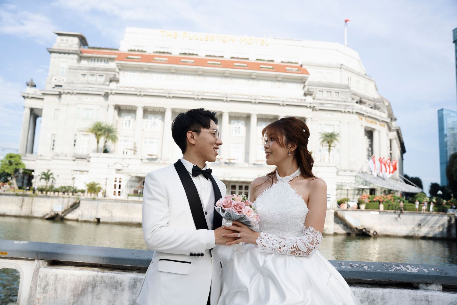 Bride in white gown holding a pink bouquet embraces a groom in white tuxedo before a grand white colonial building by the waterfront.