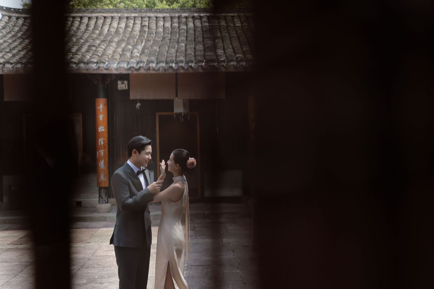 Couple in elegant attire embracing in a traditional Chinese courtyard with a tiled roof, framed by dark wooden lattice in the foreground.