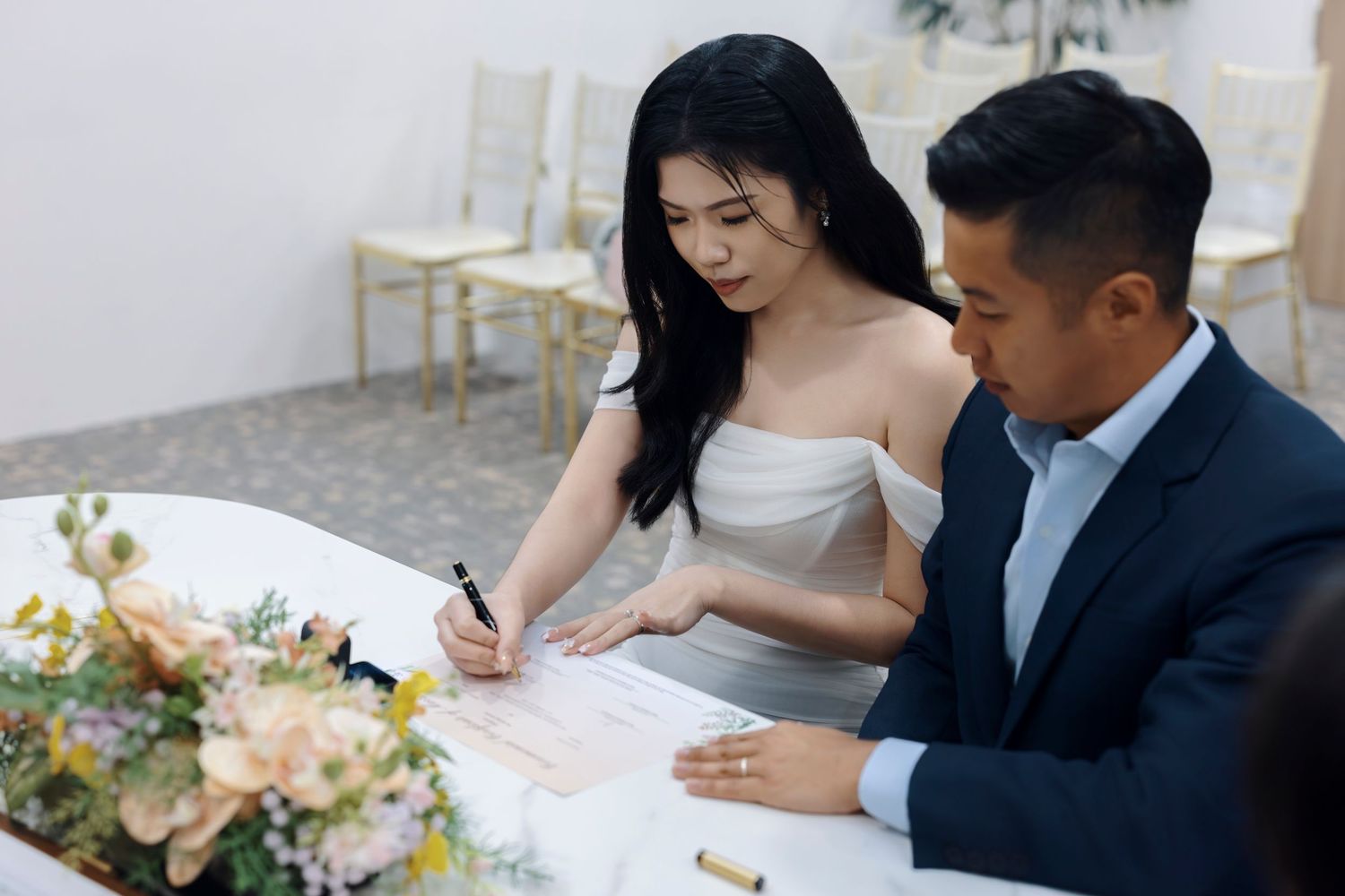 Couple signing wedding documents at a ceremony table with floral arrangements, bride in off-shoulder gown and groom in navy suit.