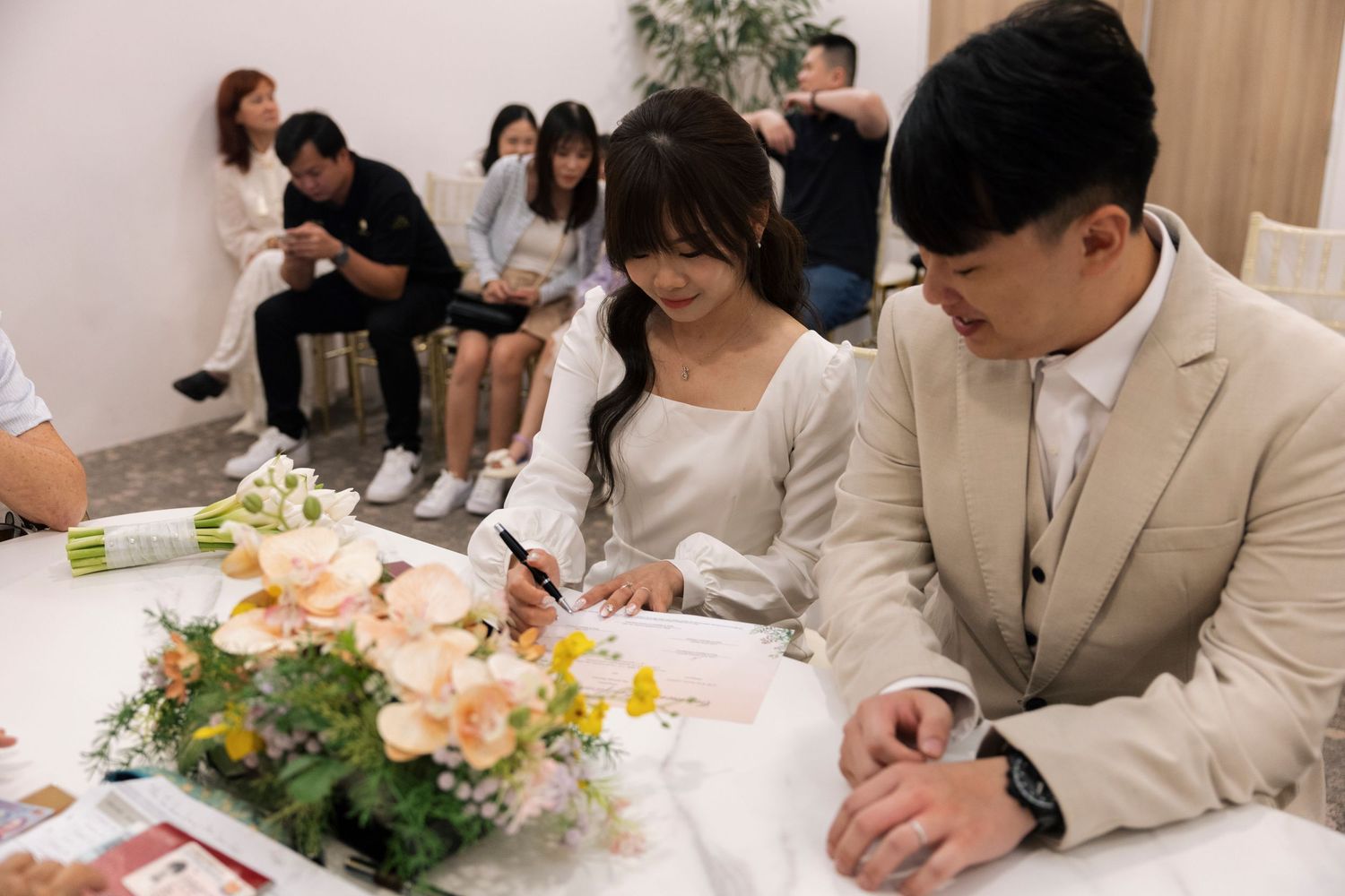 Couple signing wedding documents at a table with floral arrangement, guests seated in background.