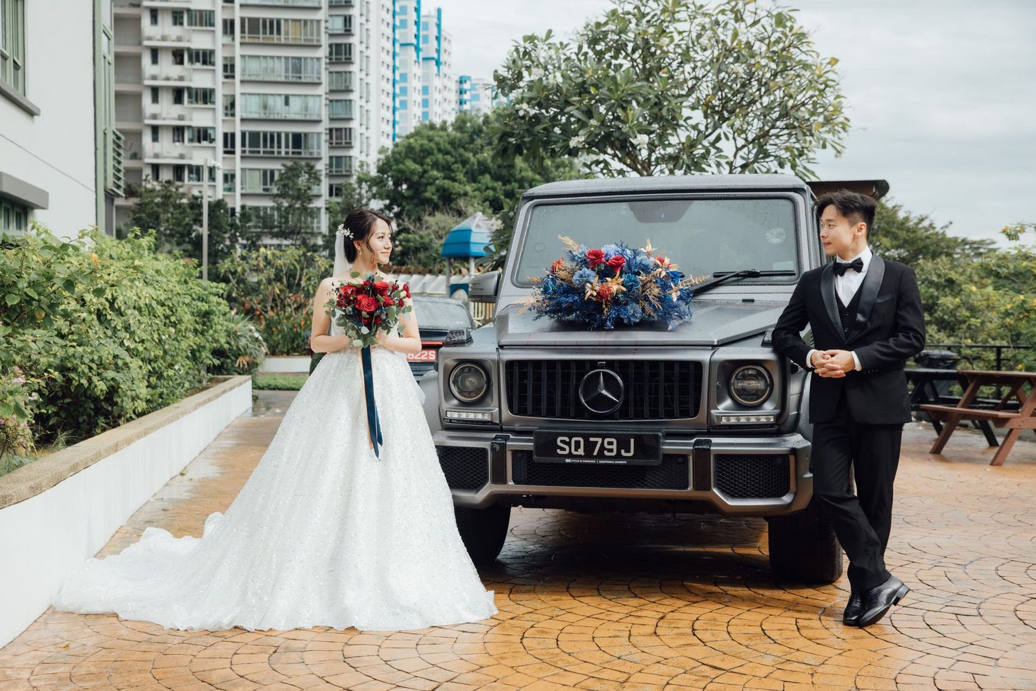 Bride in a white gown holding a bouquet and groom in a black tuxedo pose beside a Mercedes G-Class SUV decorated with flowers.