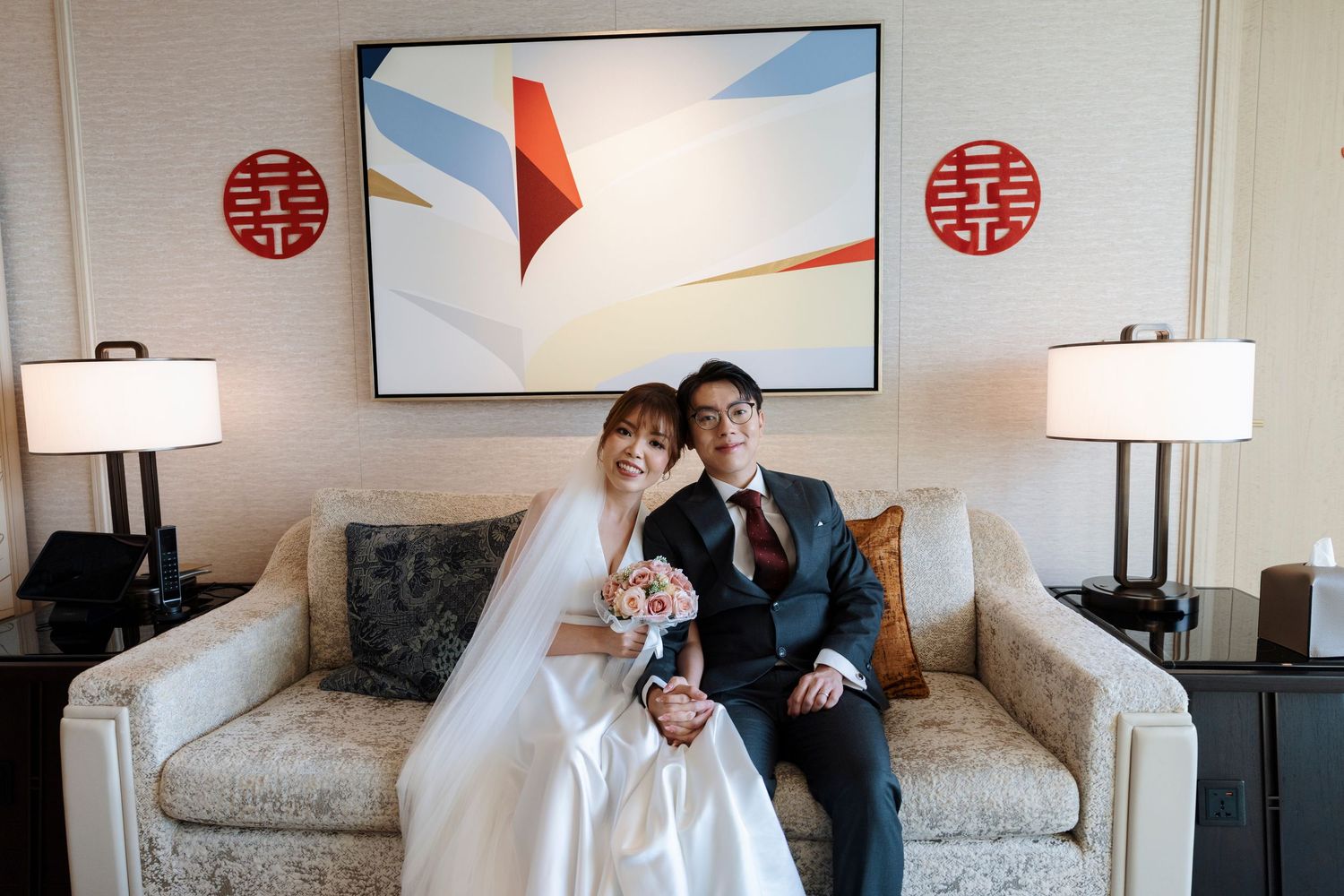 Bride in a white gown holding a pink bouquet sits beside a groom in a dark suit on a cream sofa beneath red double happiness symbols.