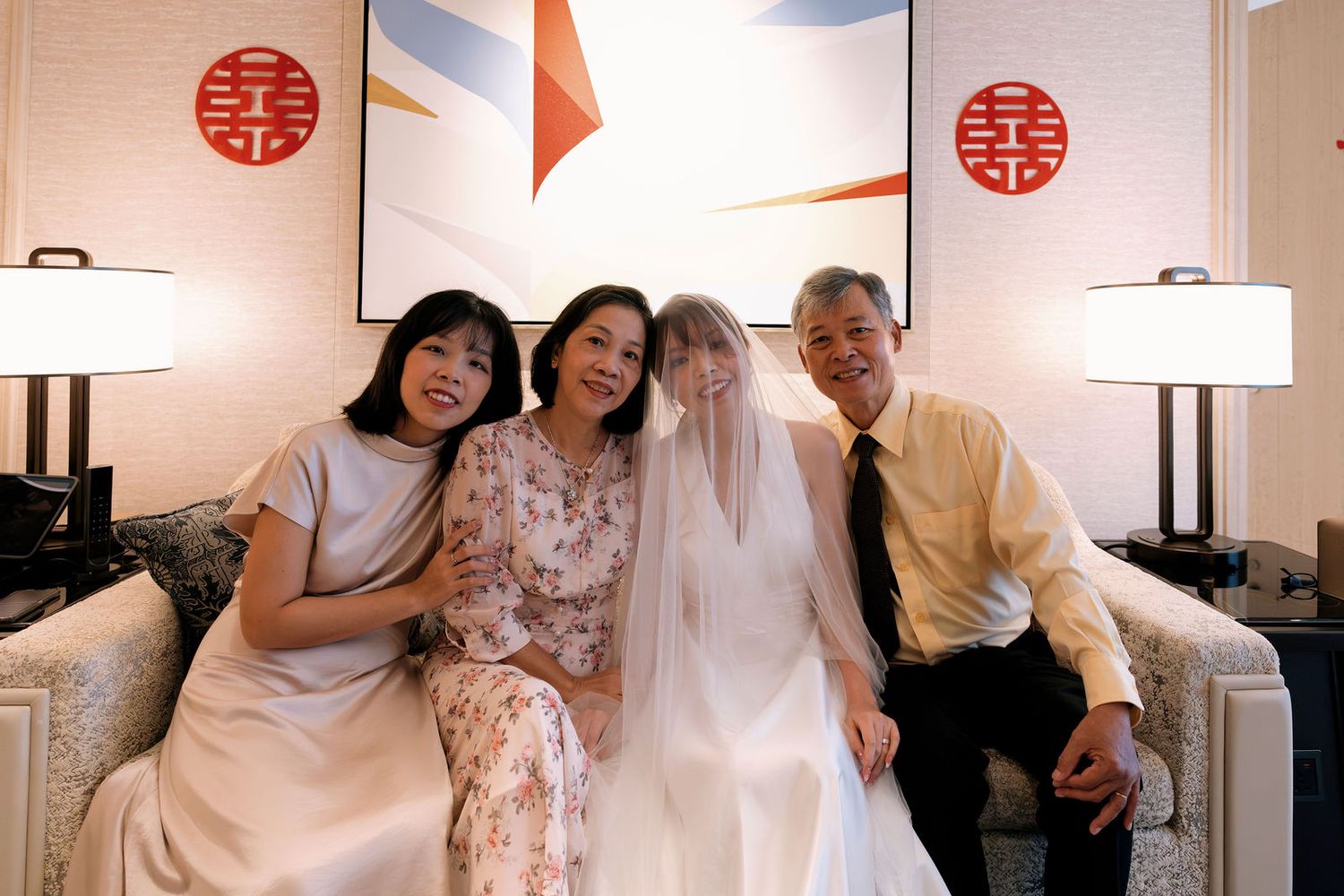 Bride in white veil poses with family on sofa, double happiness symbols on wall, wedding celebration.