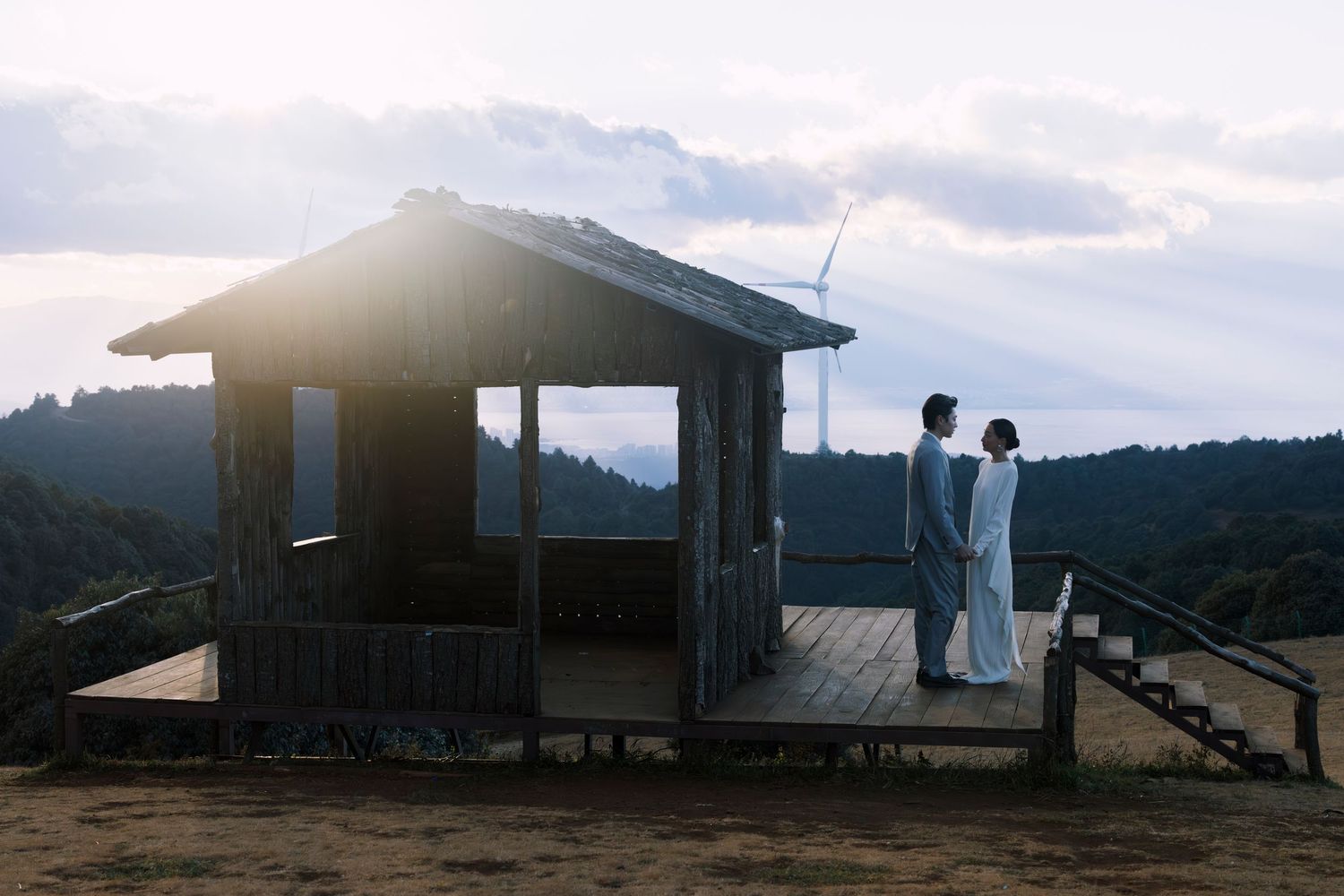Couple in matching grey outfits stand together on a wooden deck at dusk with a wind turbine behind them.