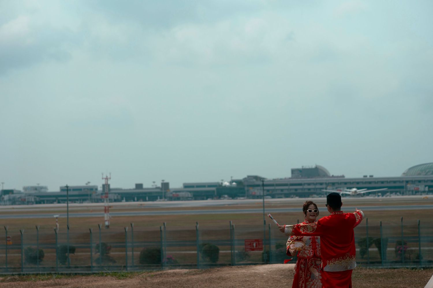 Couple in traditional red attire posing at an airport observation deck with runway and terminal in background.