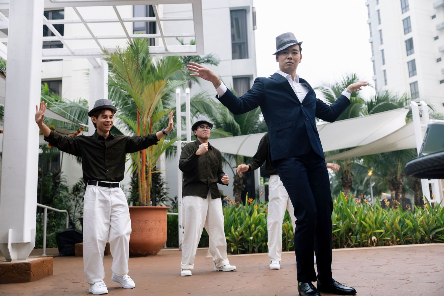 Three men in stylish hats dance outdoors near a modern building surrounded by tropical plants.