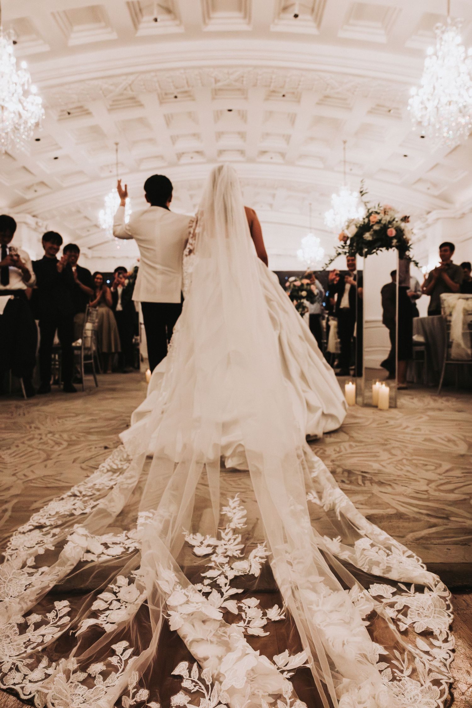 Bride in a lace wedding gown with a long floral cathedral veil walks down the aisle of an elegant ballroom with chandeliers toward guests.