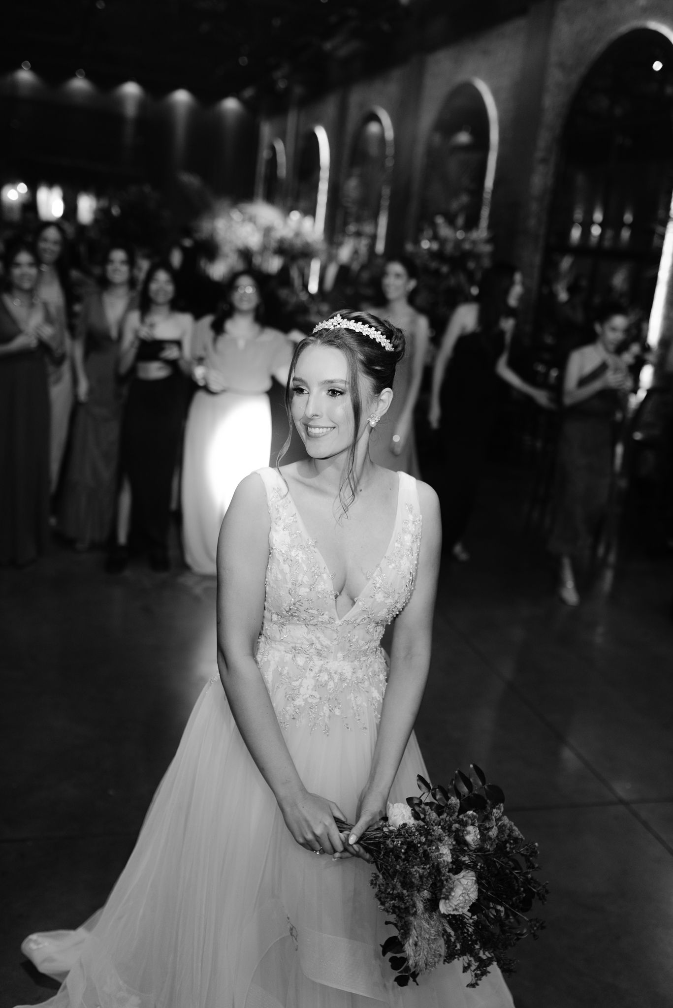 A bride in an elegant white gown holds a bouquet while standing in a dimly lit reception venue.