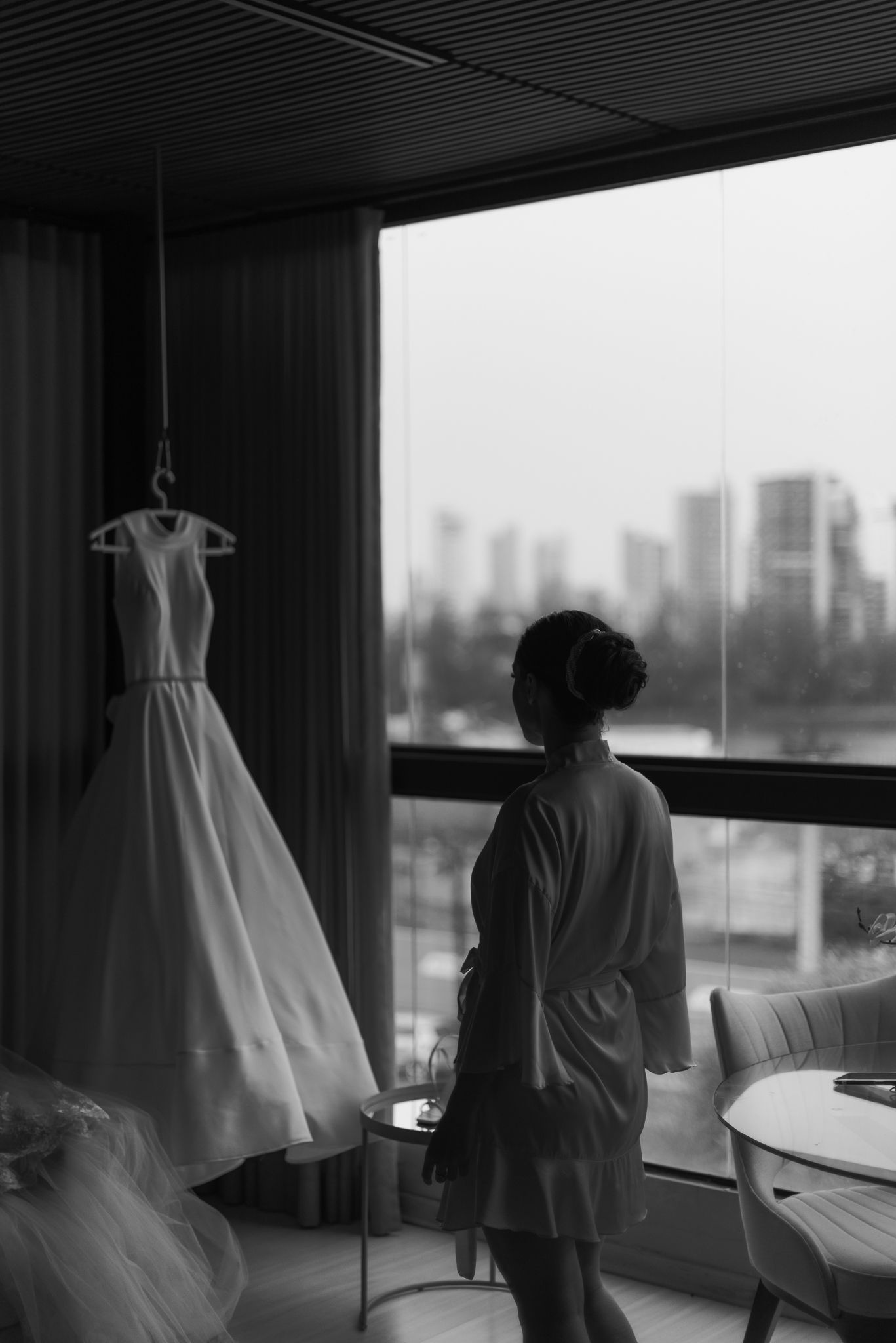 Silhouetted view of a wedding dress hanging by a window with city skyline in background.