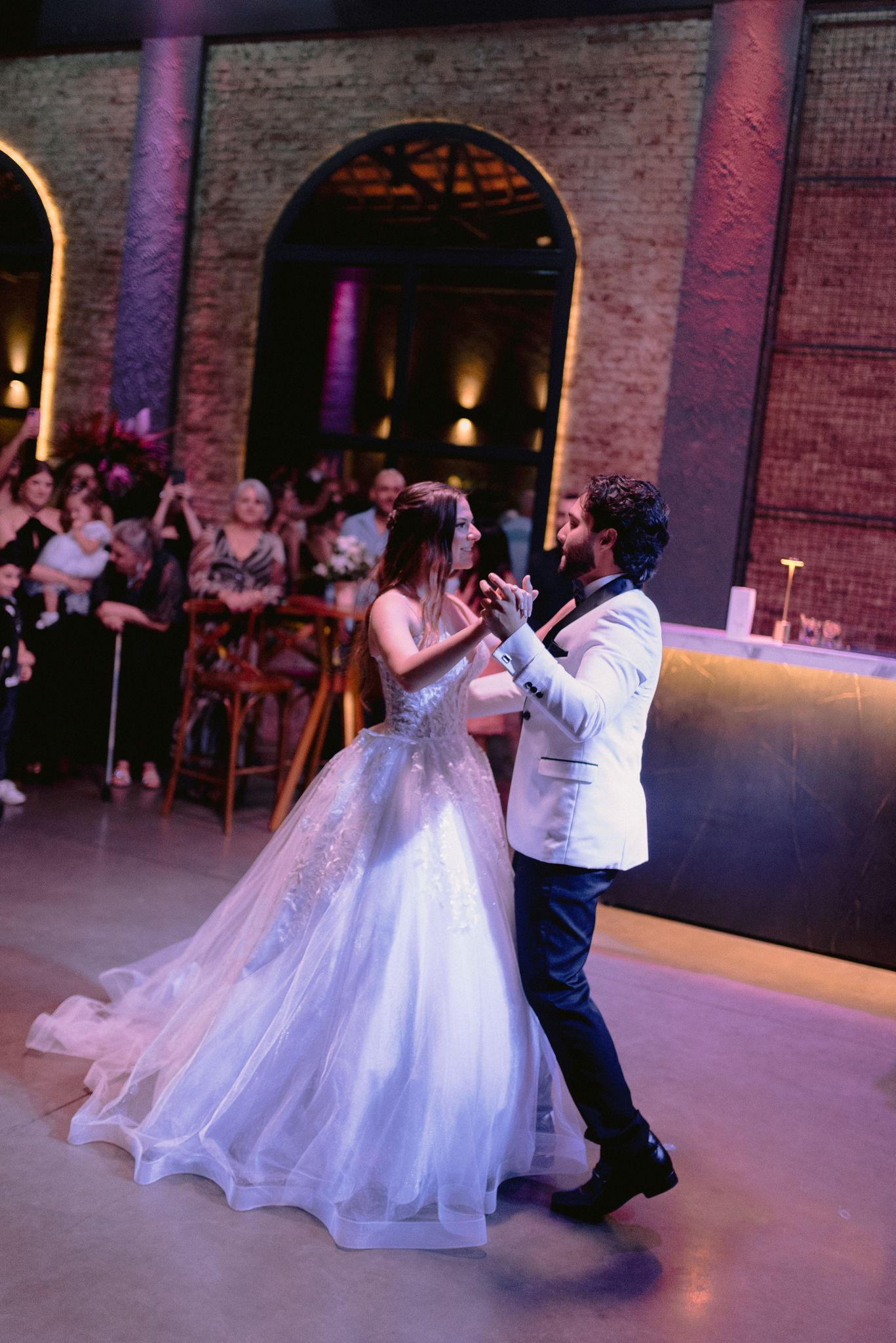 A couple performs their first dance at a wedding reception in a dimly lit industrial-style venue with brick walls.