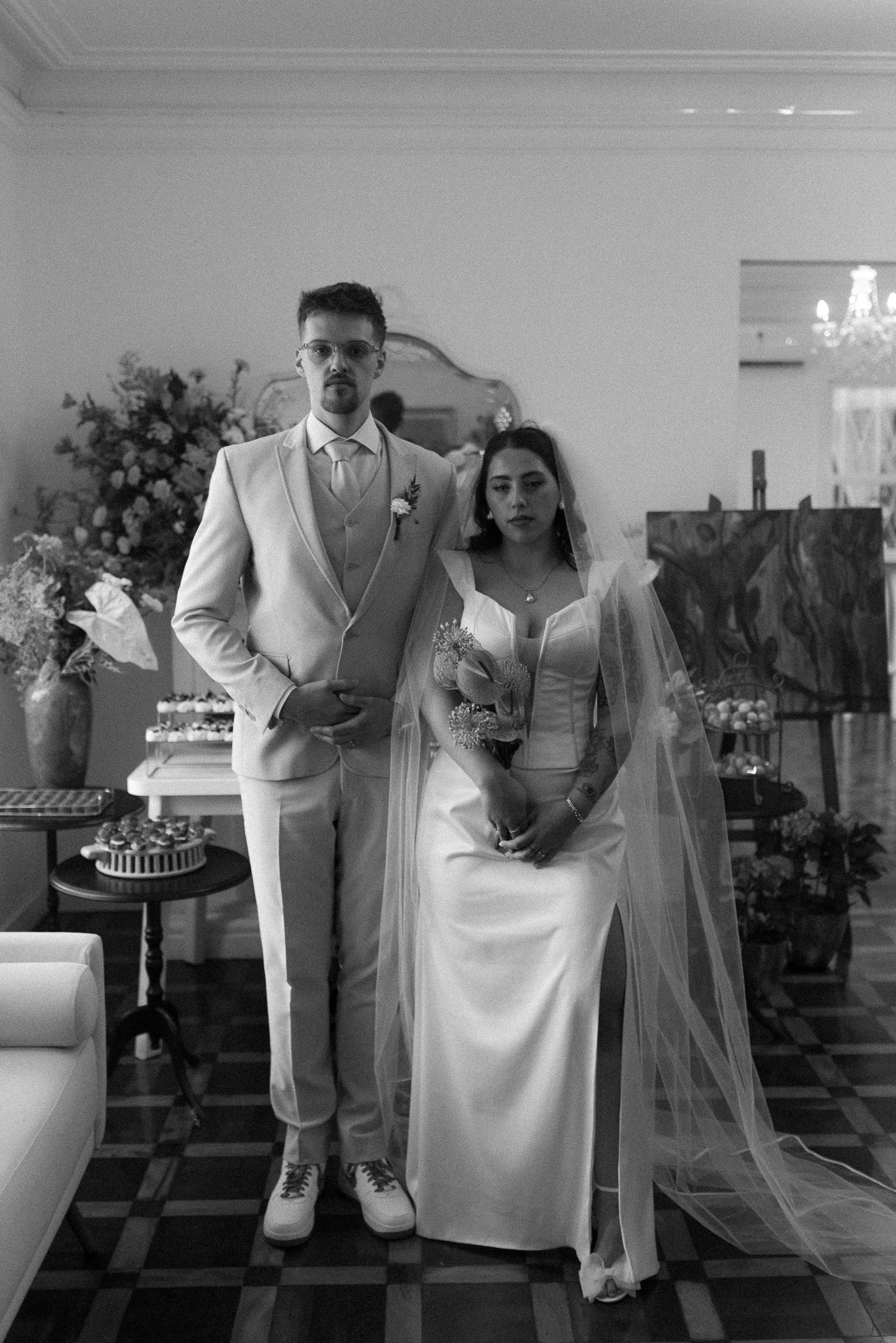 Wedding party poses in formal attire against white interior walls with checkerboard floors.