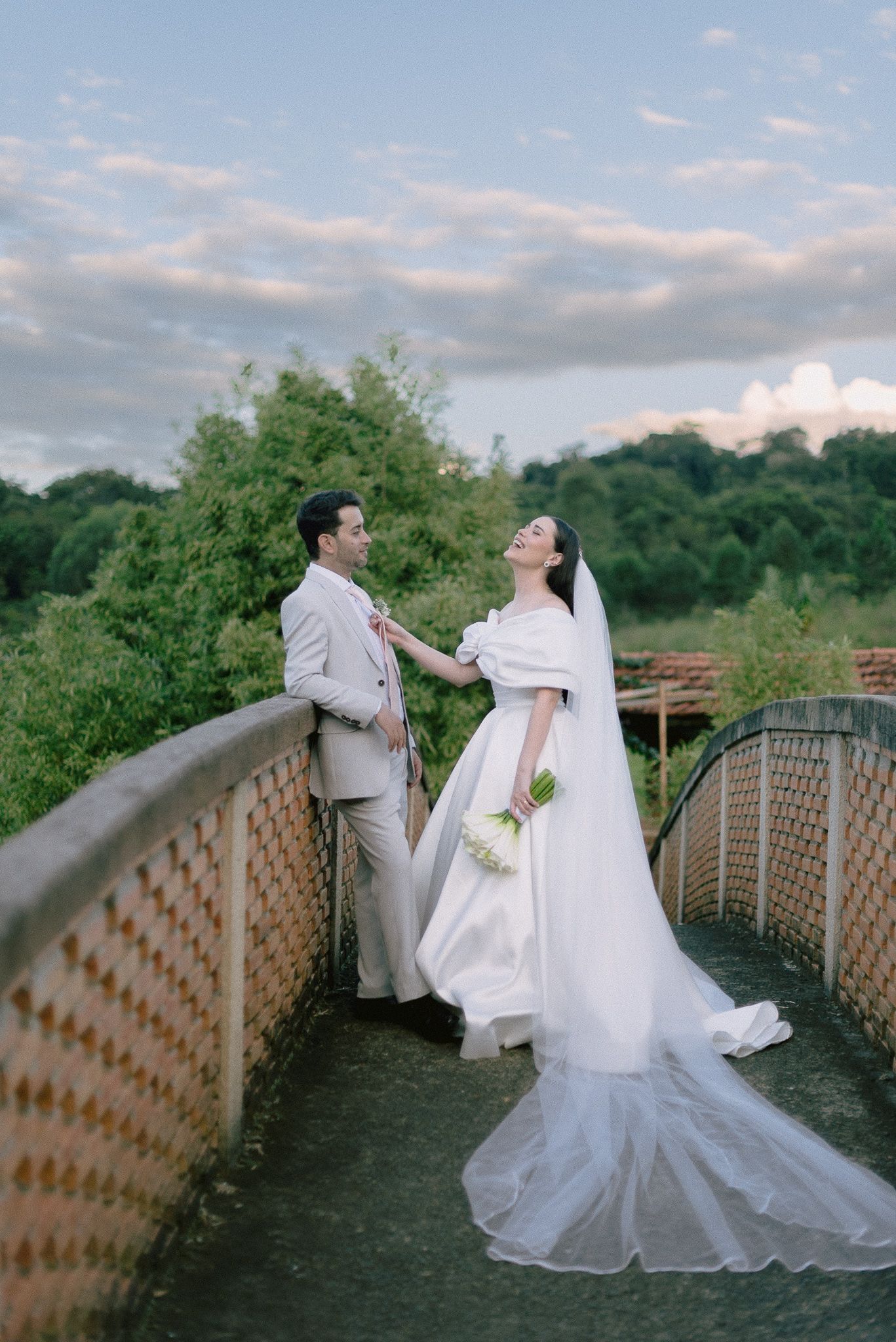 A couple in wedding attire share a romantic moment on a rustic footbridge surrounded by green foliage and clouds.