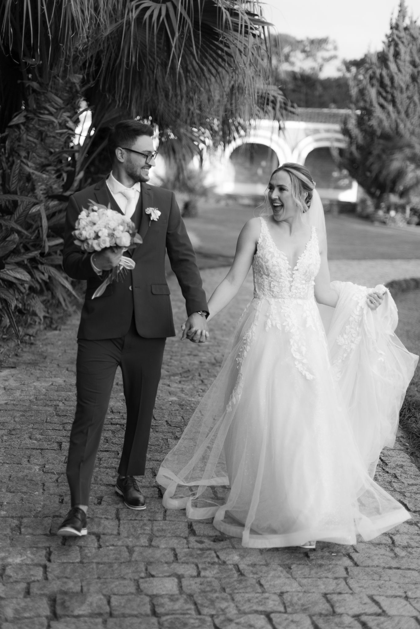 Black and white photo of newlyweds walking along a stone pathway near tropical plants.