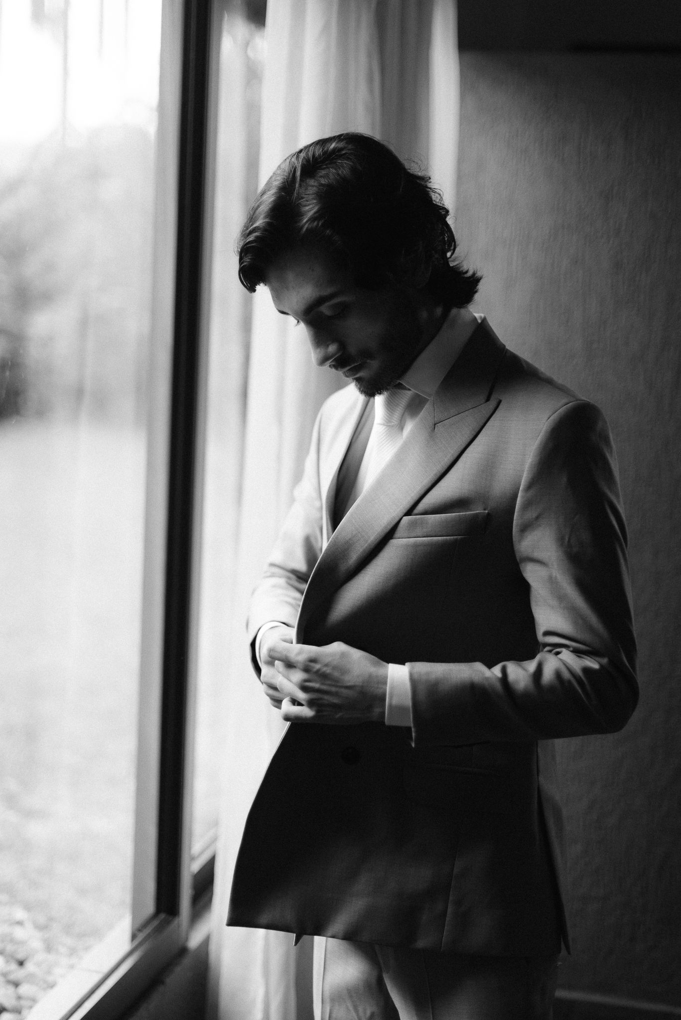 Black and white portrait of a well-dressed person in a suit jacket standing by a window in dramatic lighting.