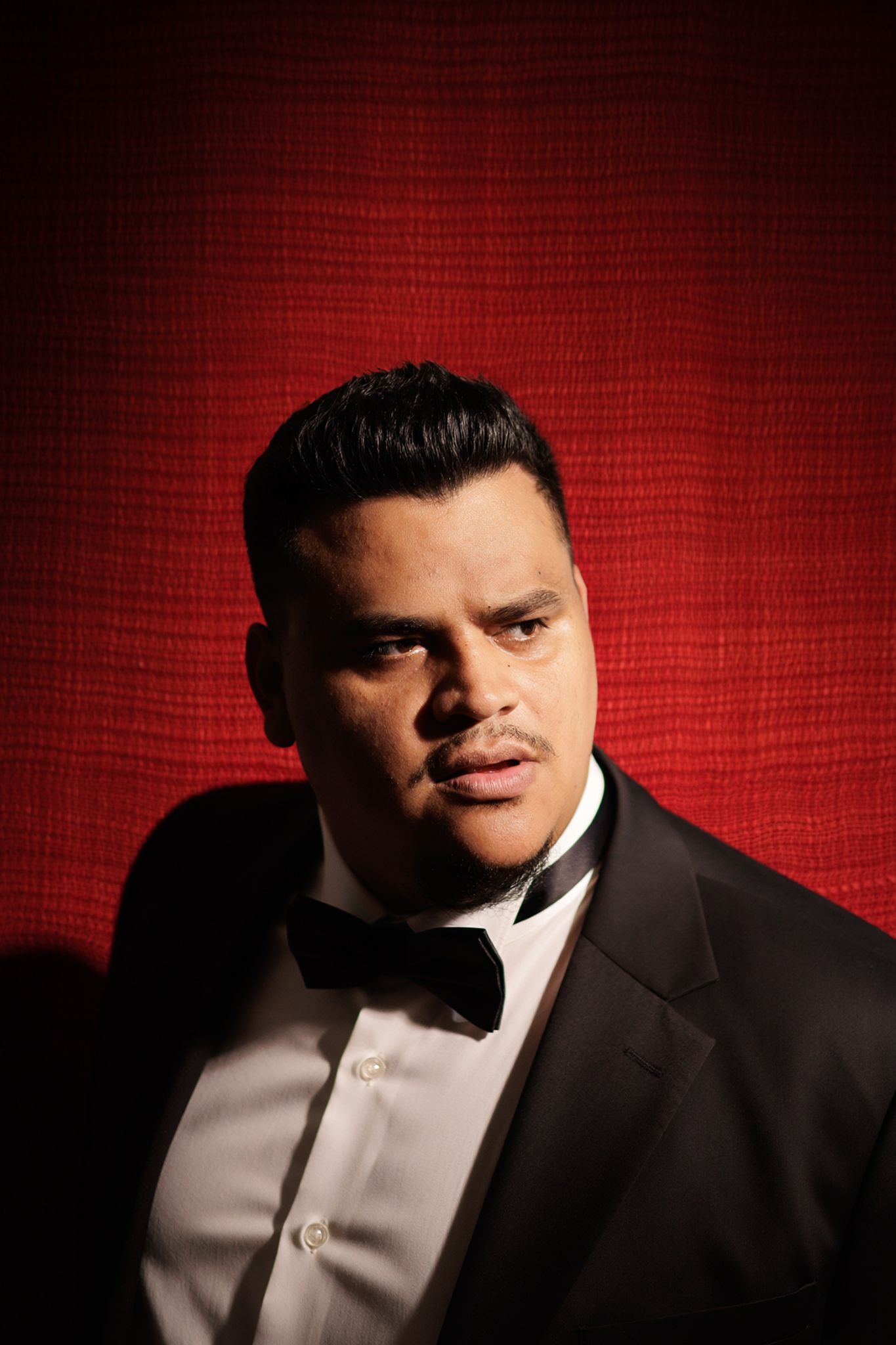 Man in tuxedo with bow tie posing against a red textured background.