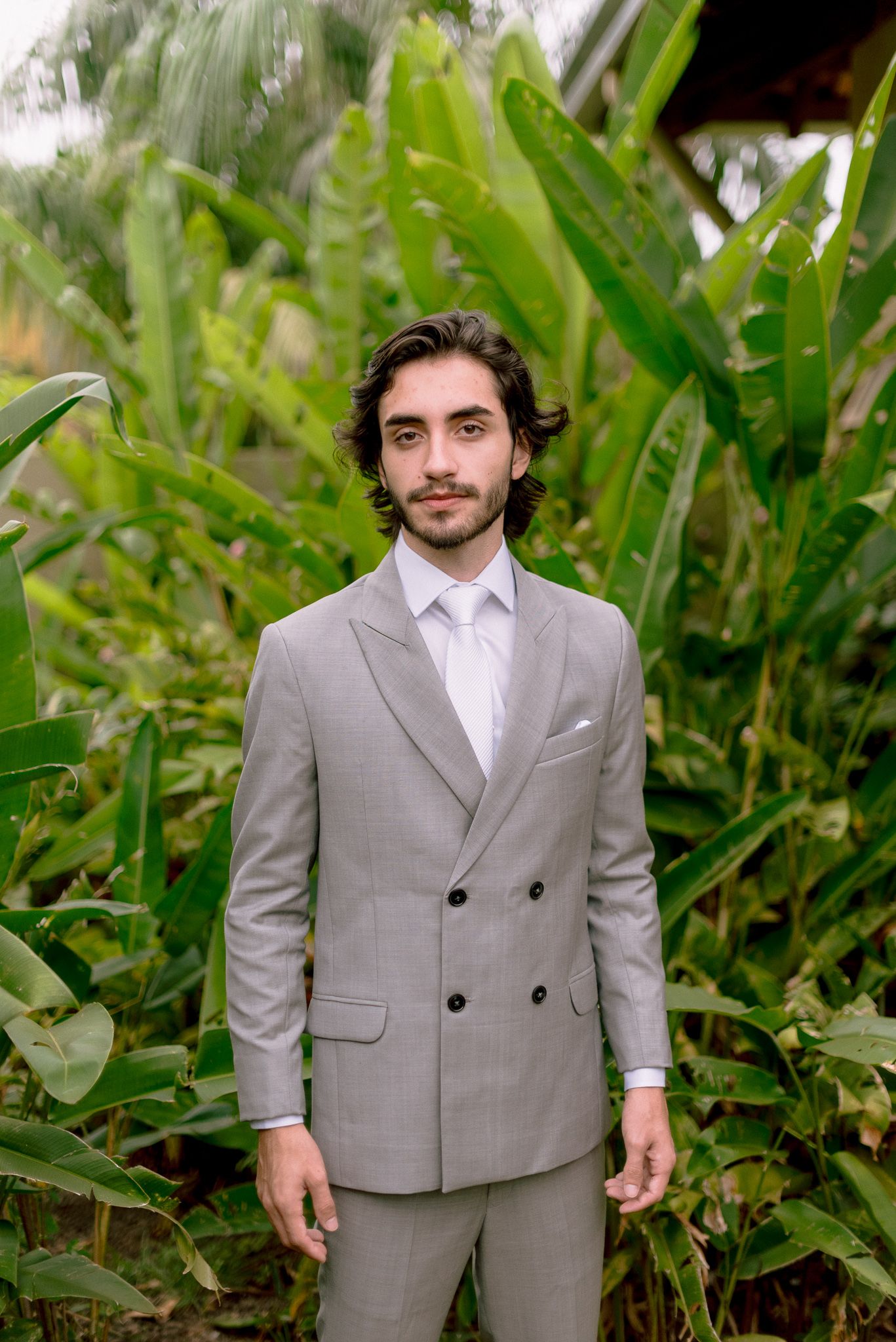 A person wearing a light gray suit stands among lush green tropical foliage in an outdoor setting.