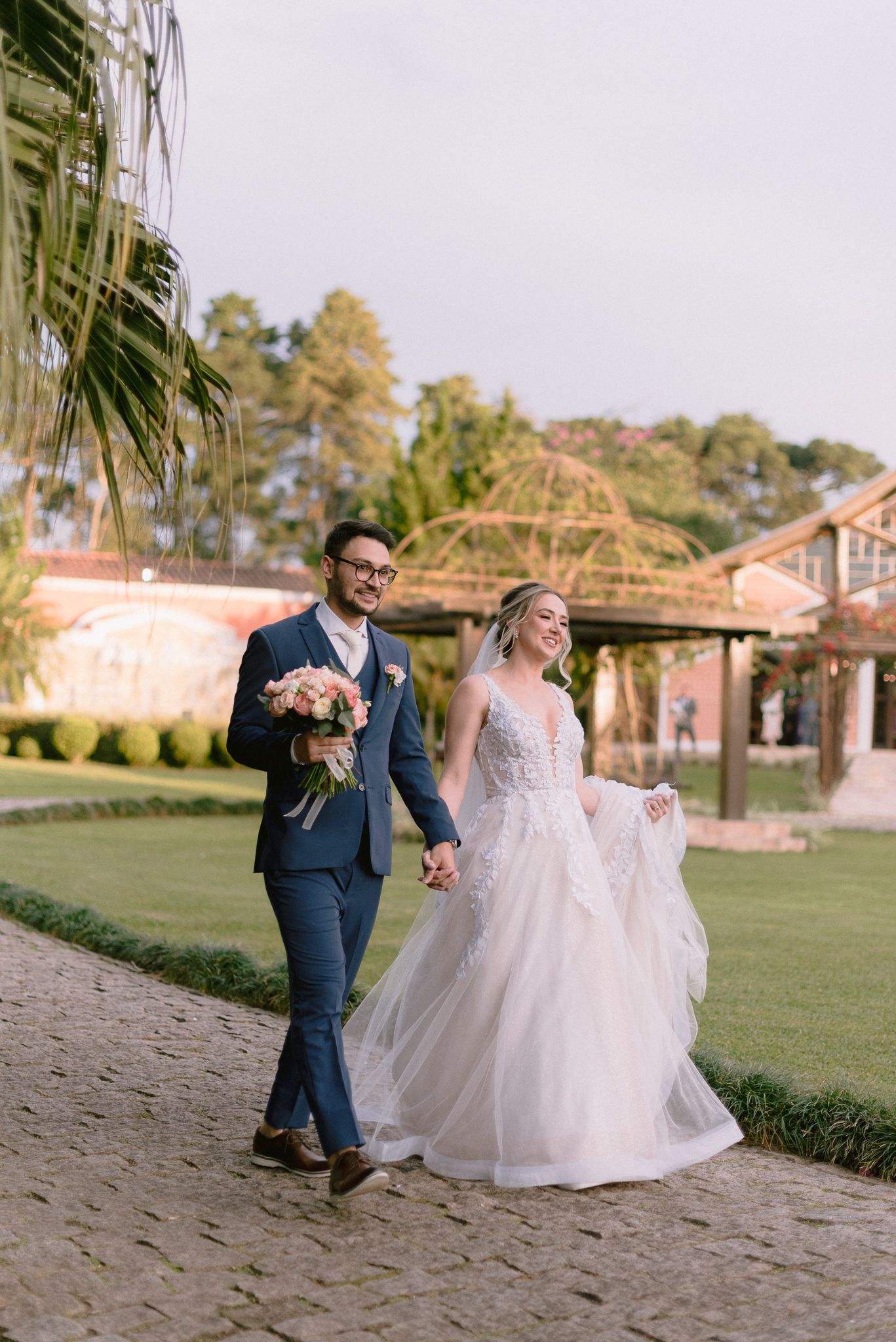 Newlyweds walking together on path holding hands with pink bouquet and scenic background.