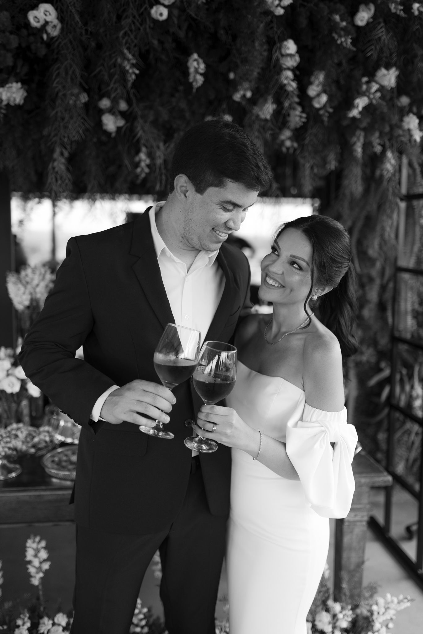 Elegant black and white portrait of couple sharing drinks under floral decor.