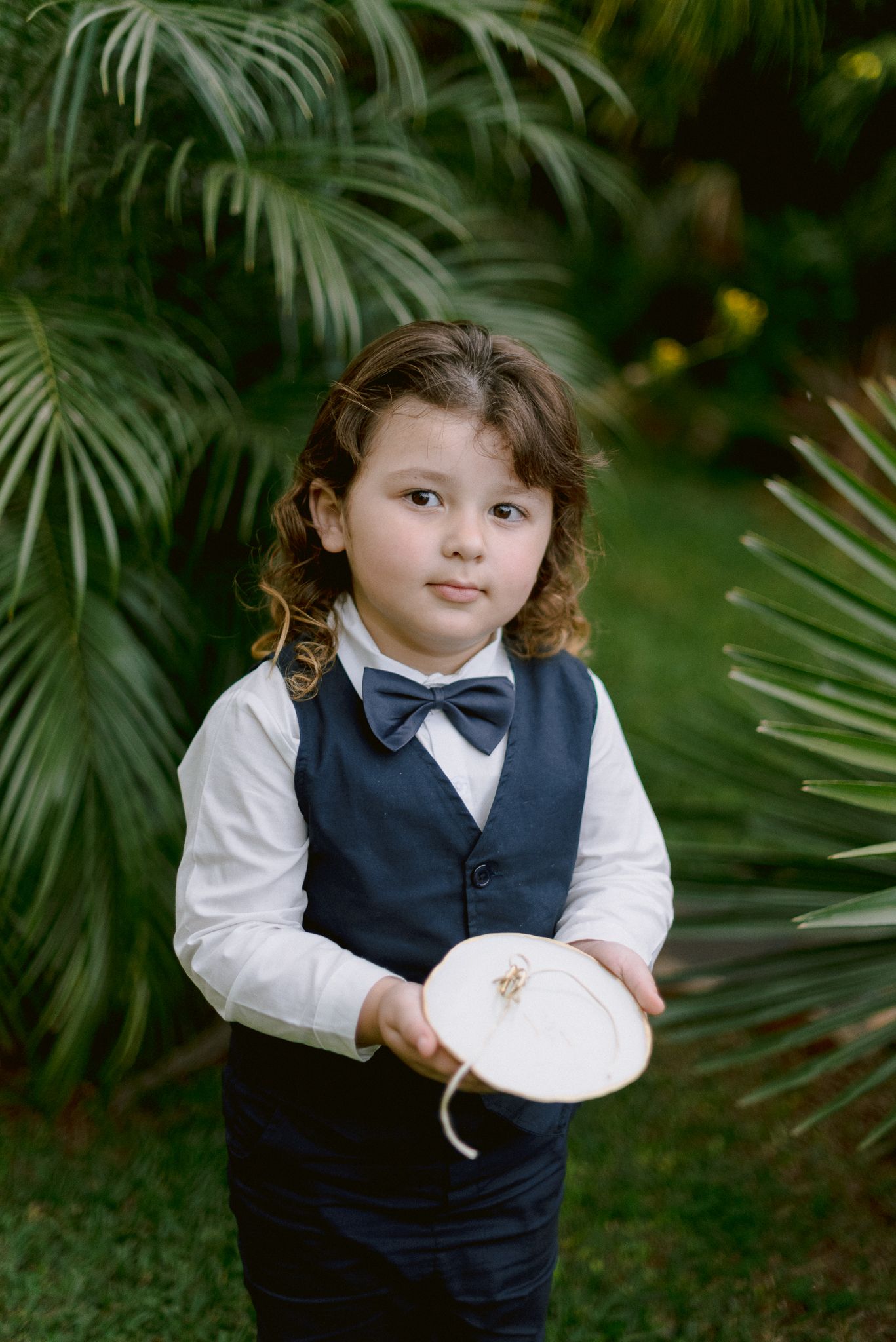 A young child in formal attire poses playfully in a garden setting with palm fronds in the background.