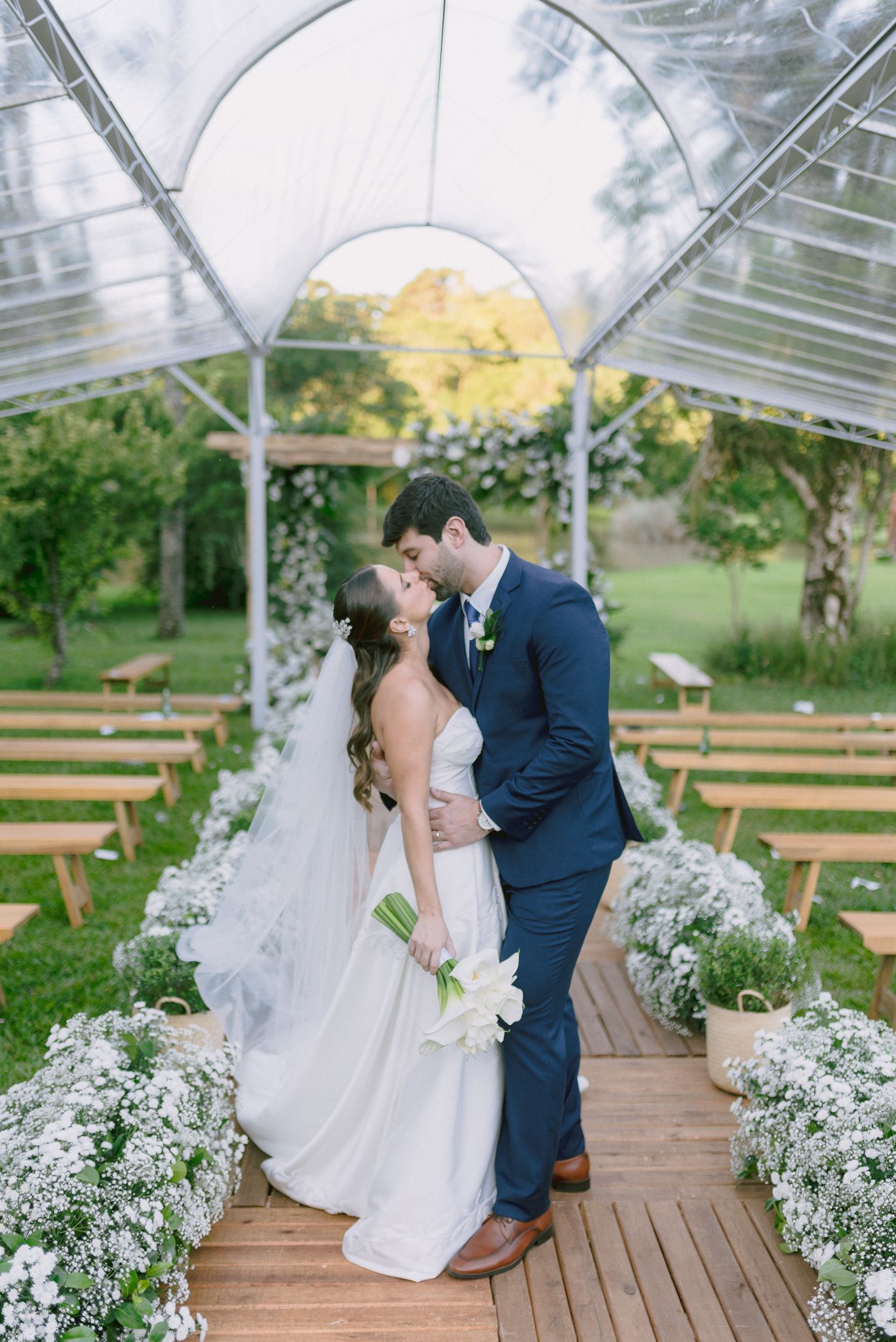 Newlyweds share a romantic kiss under a clear tent decorated with white flowers and wooden benches.